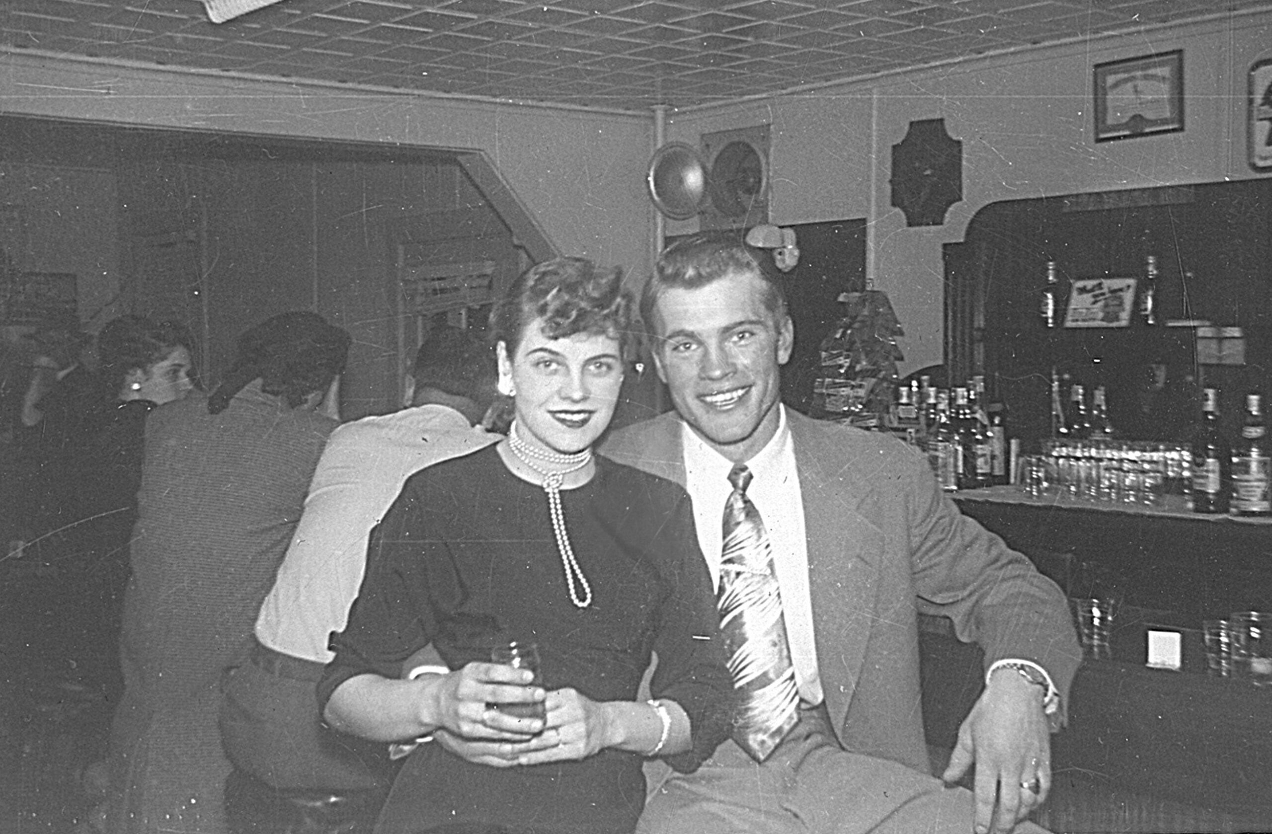 Smiling couple in vintage attire at a bar, the woman holding a drink, black-and-white photo.