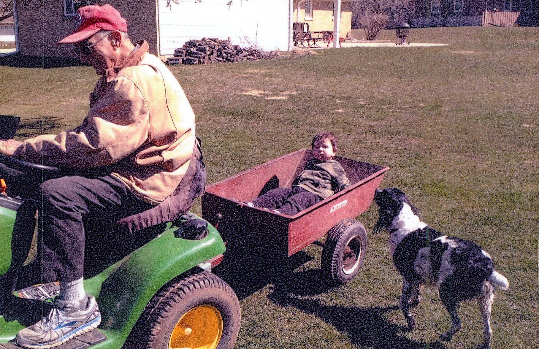 Man on a green riding lawn mower pulls a red wagon with a small child inside, while two dogs approach on the grass.
