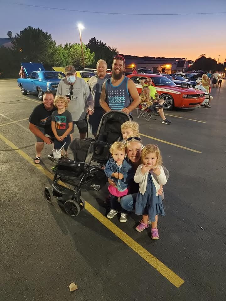 Group of adults and children posing with a stroller in a parking lot at a car show during sunset, with vintage cars in the background.