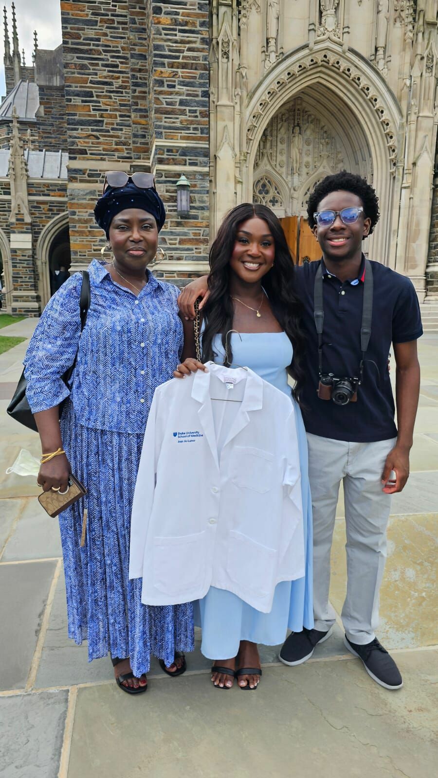 Three people pose outside a stone cathedral, center woman in pale blue dress holding a white lab coat while smiling with two friends.
