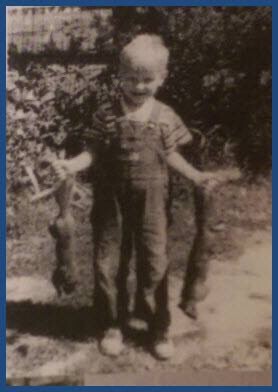 Black-and-white photo of two young children outdoors; the older child in front smiles, holding a long object in each hand, while a smaller child stands behind.