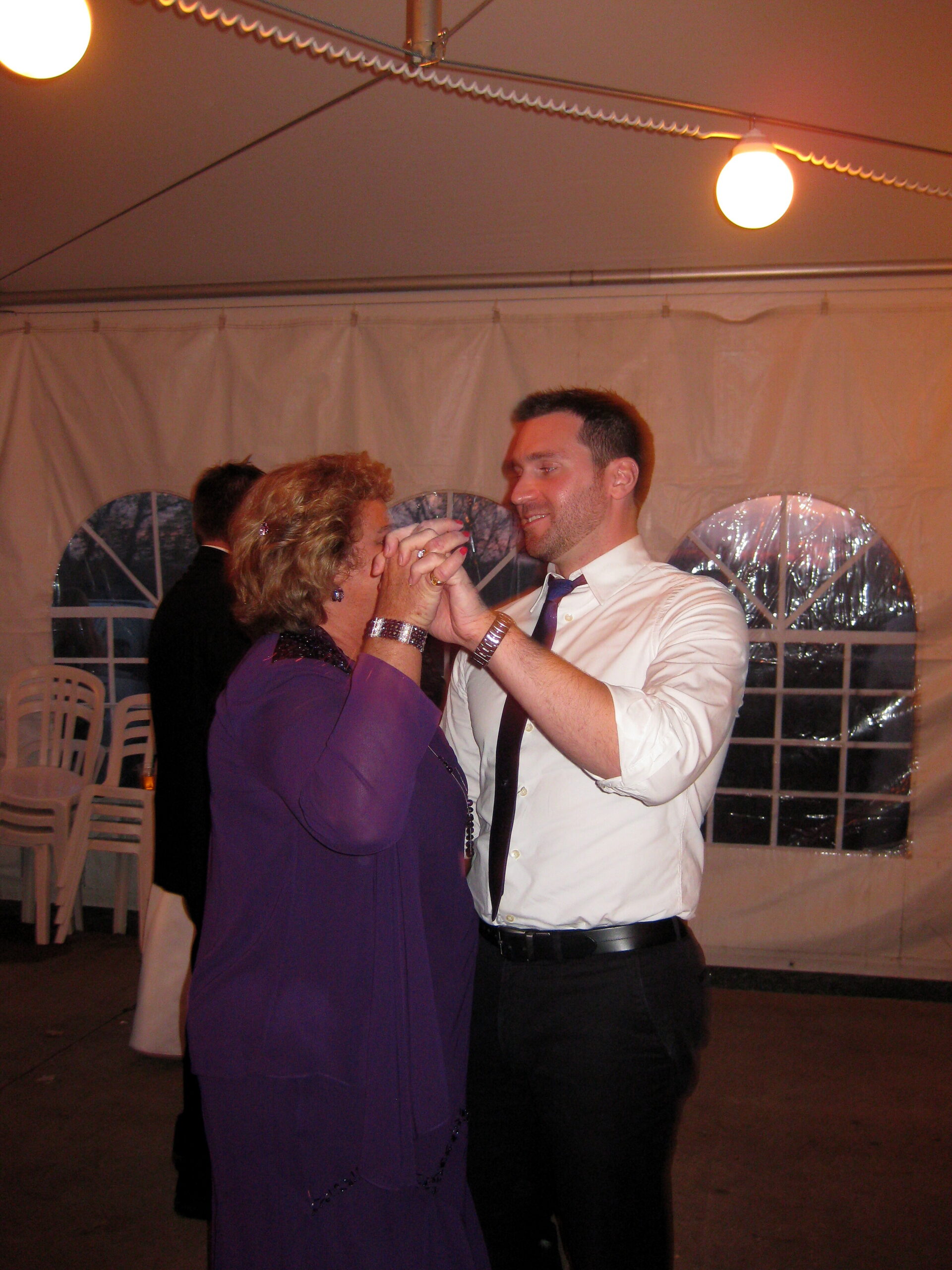 Man in a white shirt and tie dancing with an older woman in a purple dress inside a white event tent with string lights.