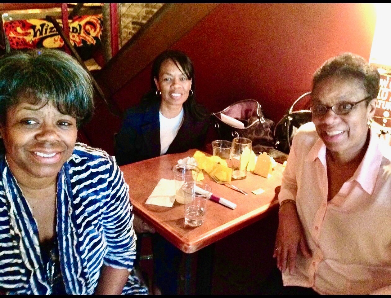 Three women sit around a restaurant table smiling for a selfie; yellow napkins and glasses on the table visible in the foreground and background.