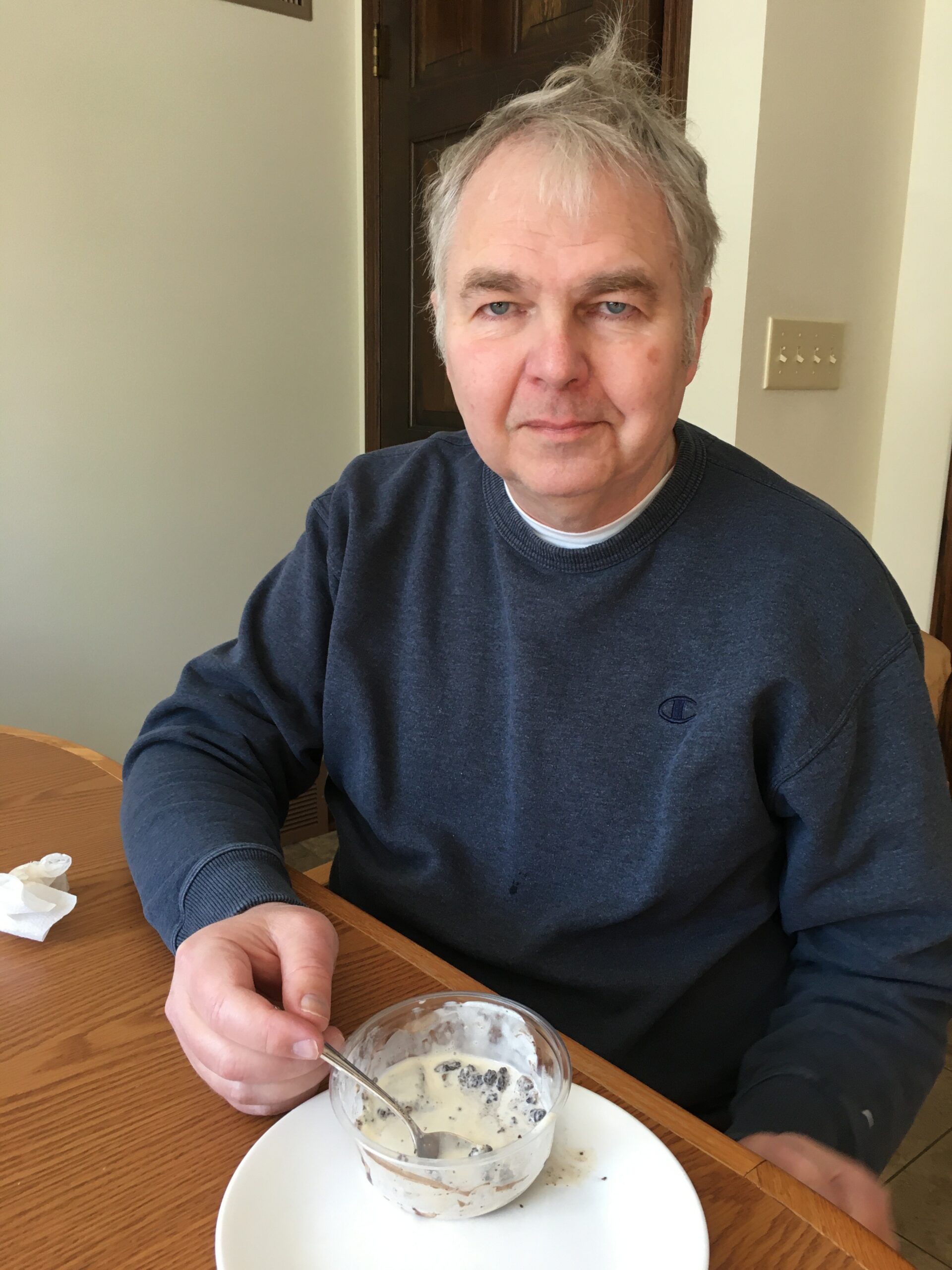 Man in a dark blue sweatshirt sits at a wooden table, reaching for a spoon next to a cup of cookies and cream ice cream dessert.