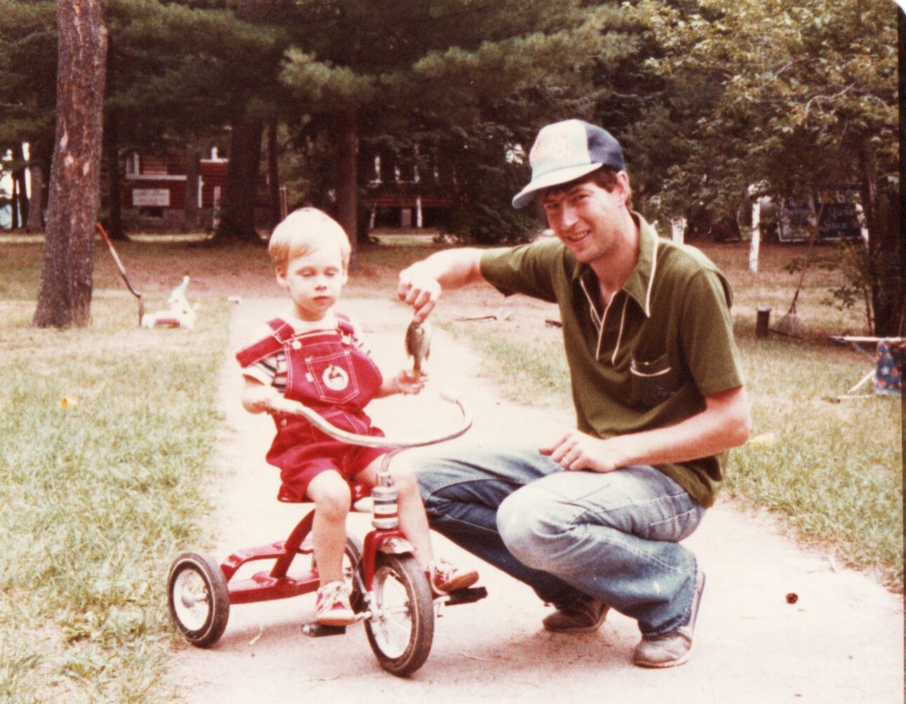 Man kneeling beside a toddler on a red tricycle in a park, holding up a small fish for him to see.