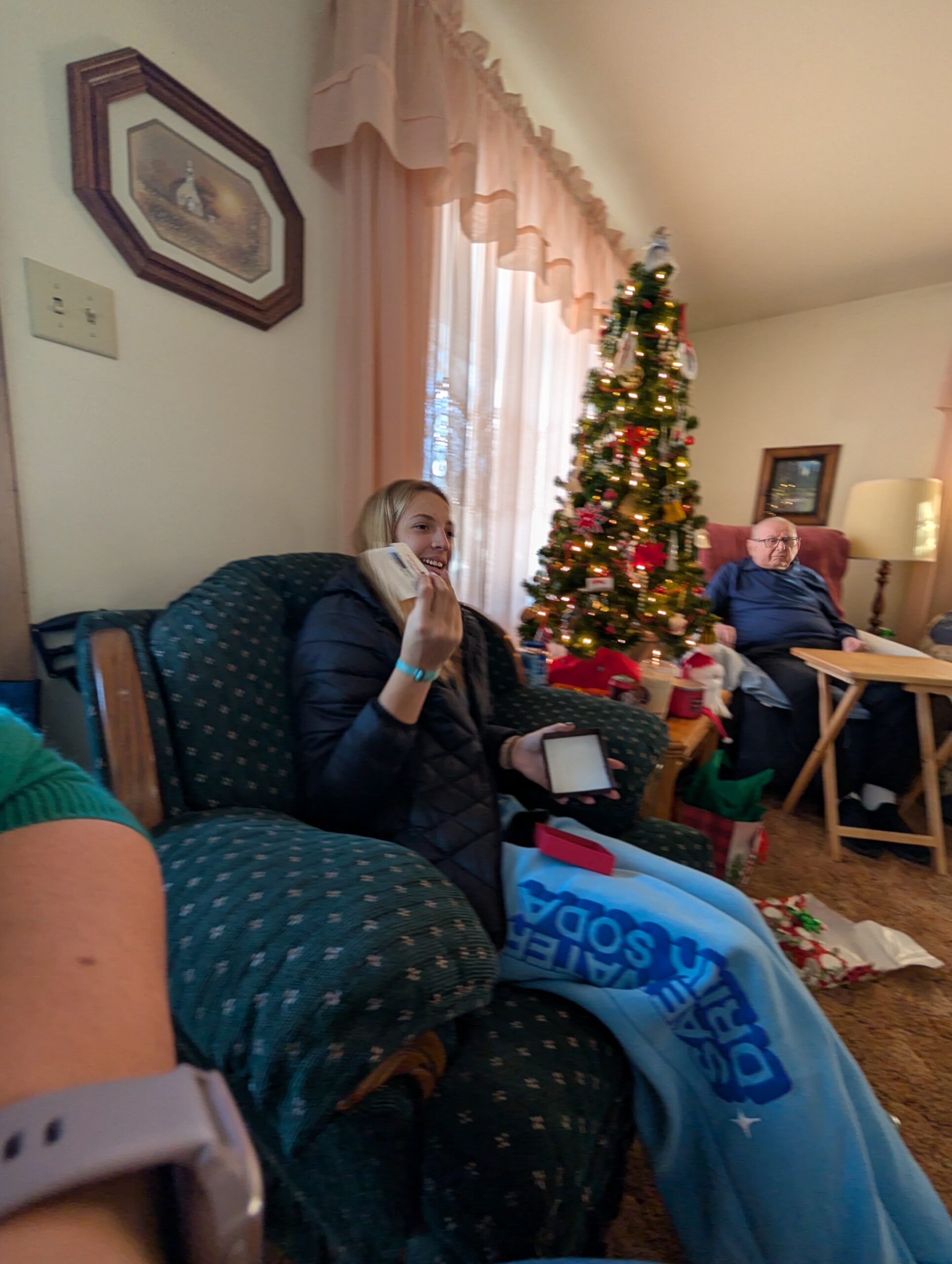 Living room during Christmas: a decorated tree by the window, a woman in a dark jacket seated and smiling, a man in a blue shirt on a chair in the background, gifts on the floor.