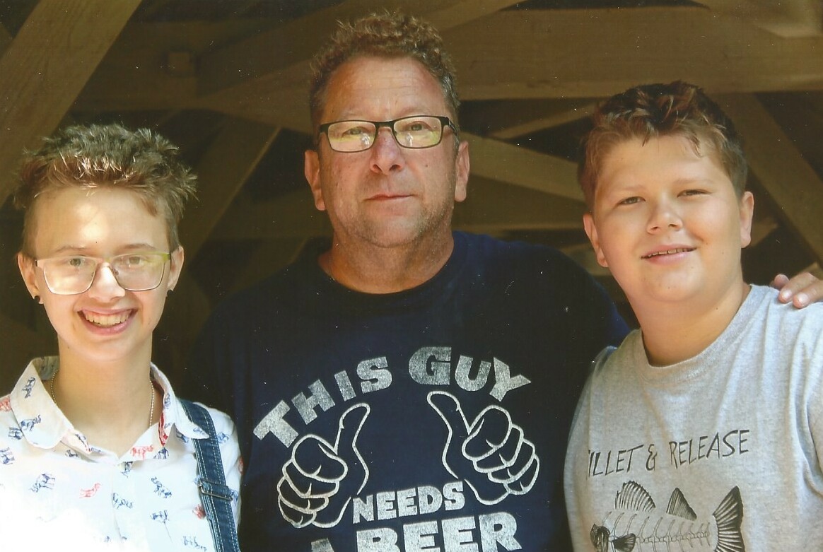 Three people posing together for a photo: a young person with glasses and a patterned shirt on the left, a man in the middle wearing a dark T-shirt, and a smiling teen in a gray shirt on the right.