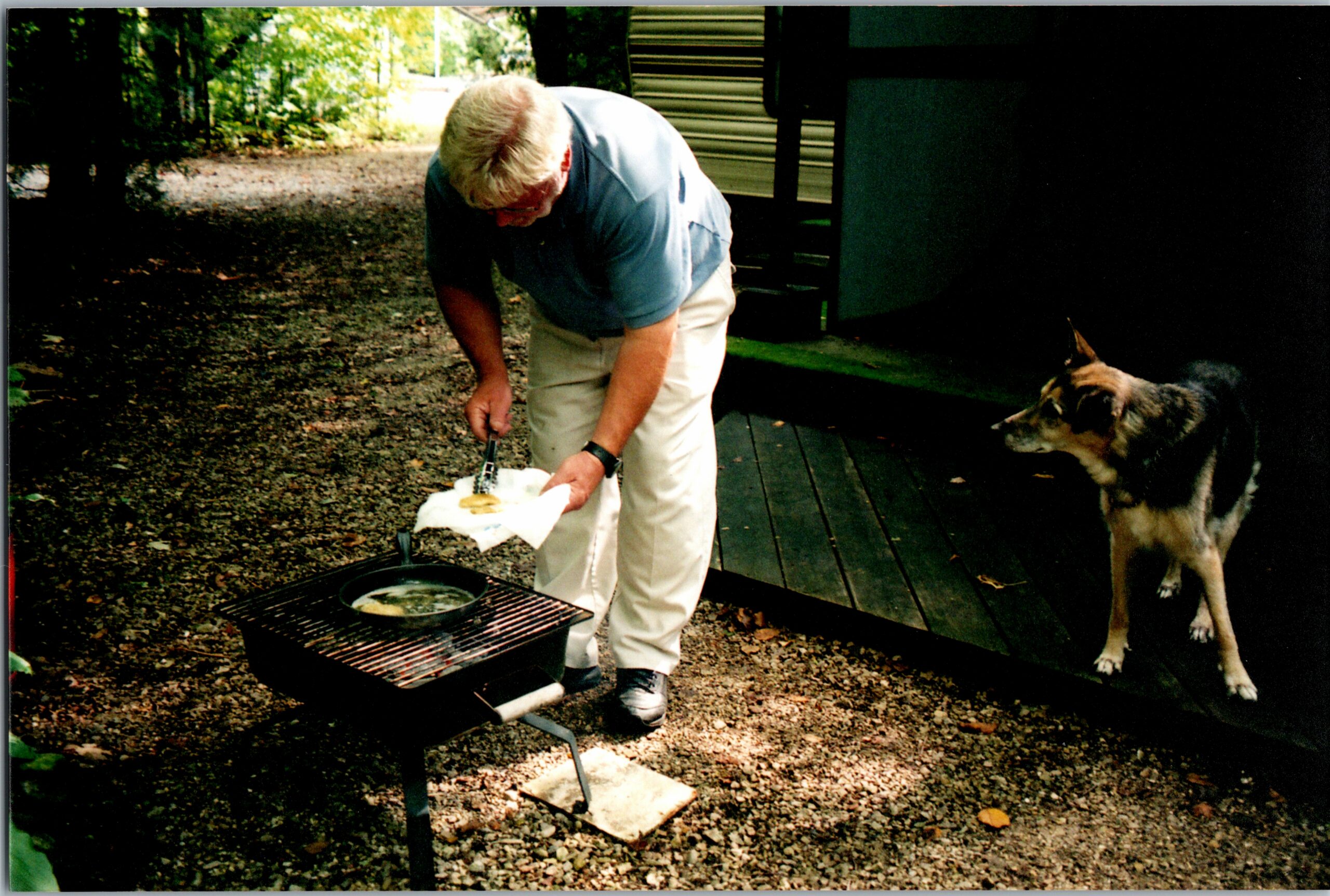 Older man bending over a grill outdoors, with a dog watching nearby