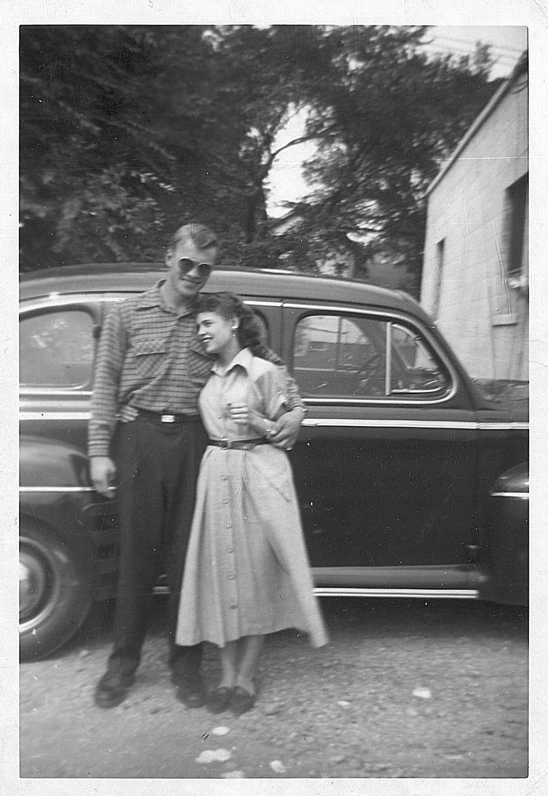 Man in sunglasses and a striped shirt stands beside a woman in a long dress in front of a vintage car outdoors, smiling together.