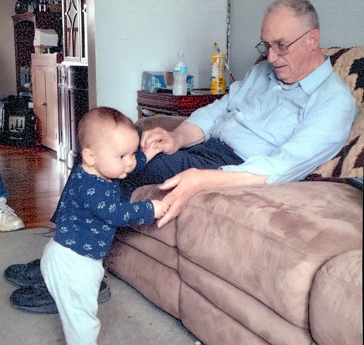 Grandfather seated on a brown couch helping a toddler stand and reach his hands in a cozy living room.