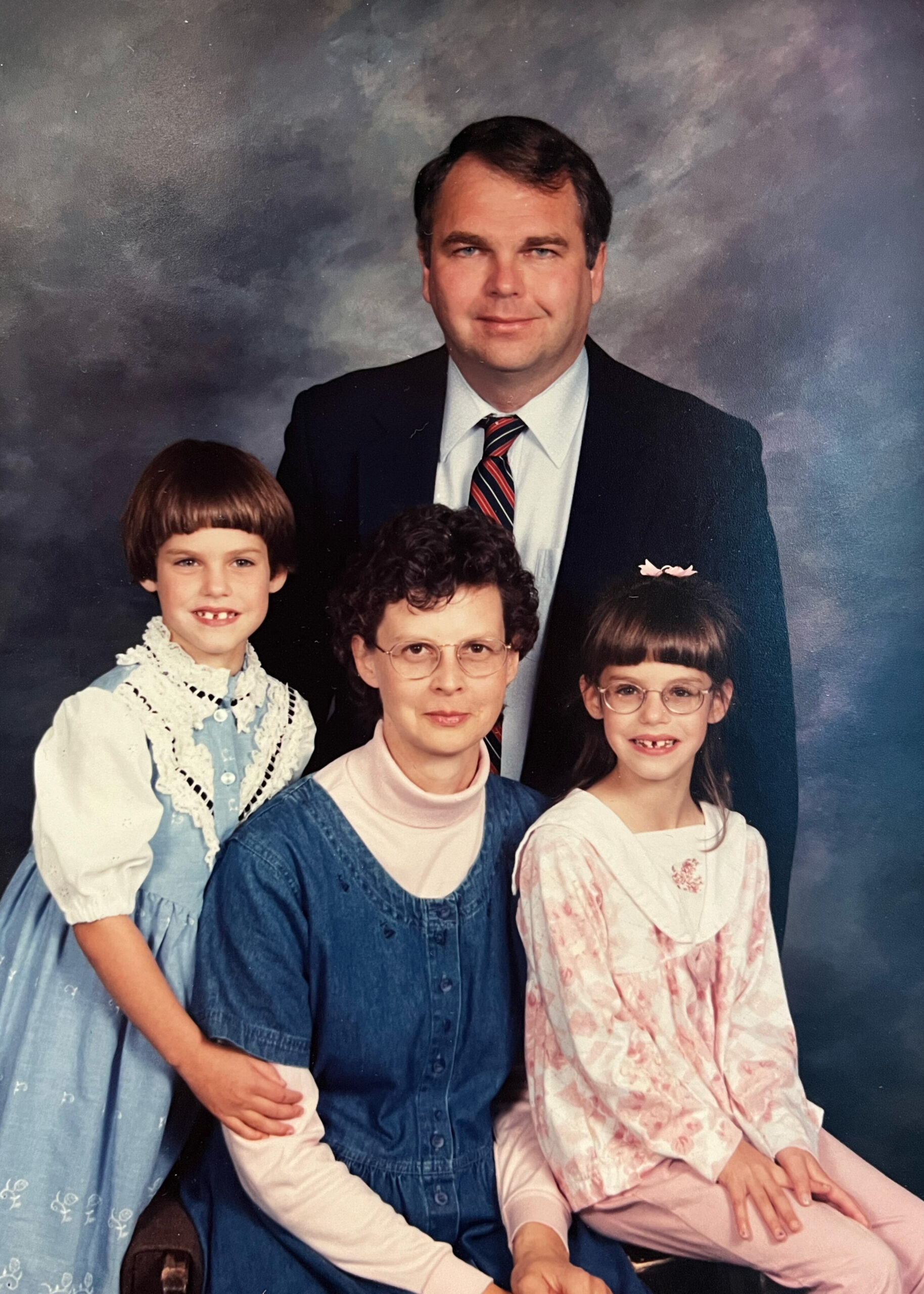 Family portrait: man in a suit stands behind a woman wearing glasses and three children in vintage clothing smiling at the camera.
