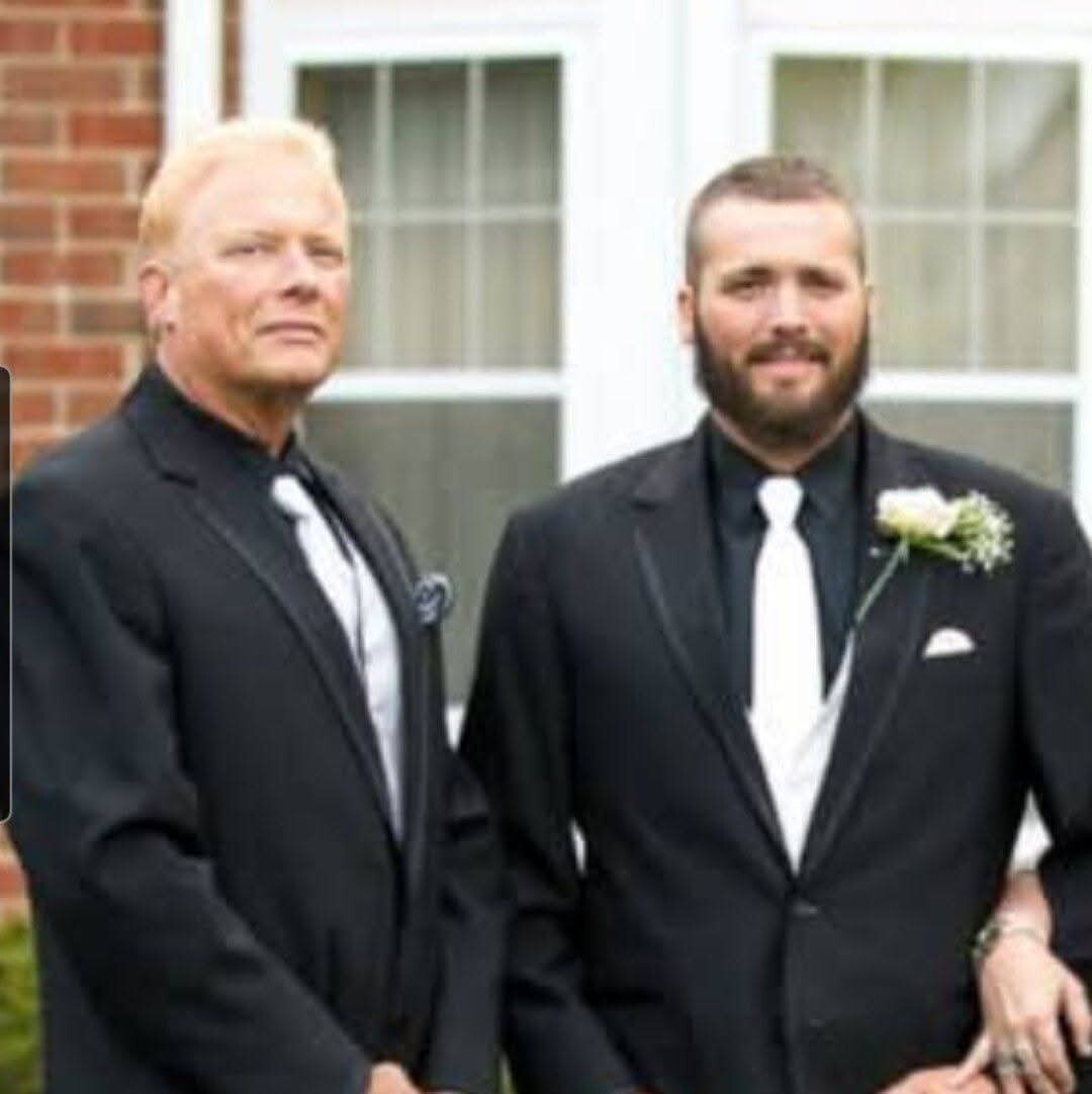 Two men in black suits with white ties standing outside in front of a brick building; one has a boutonniere on the lapel.