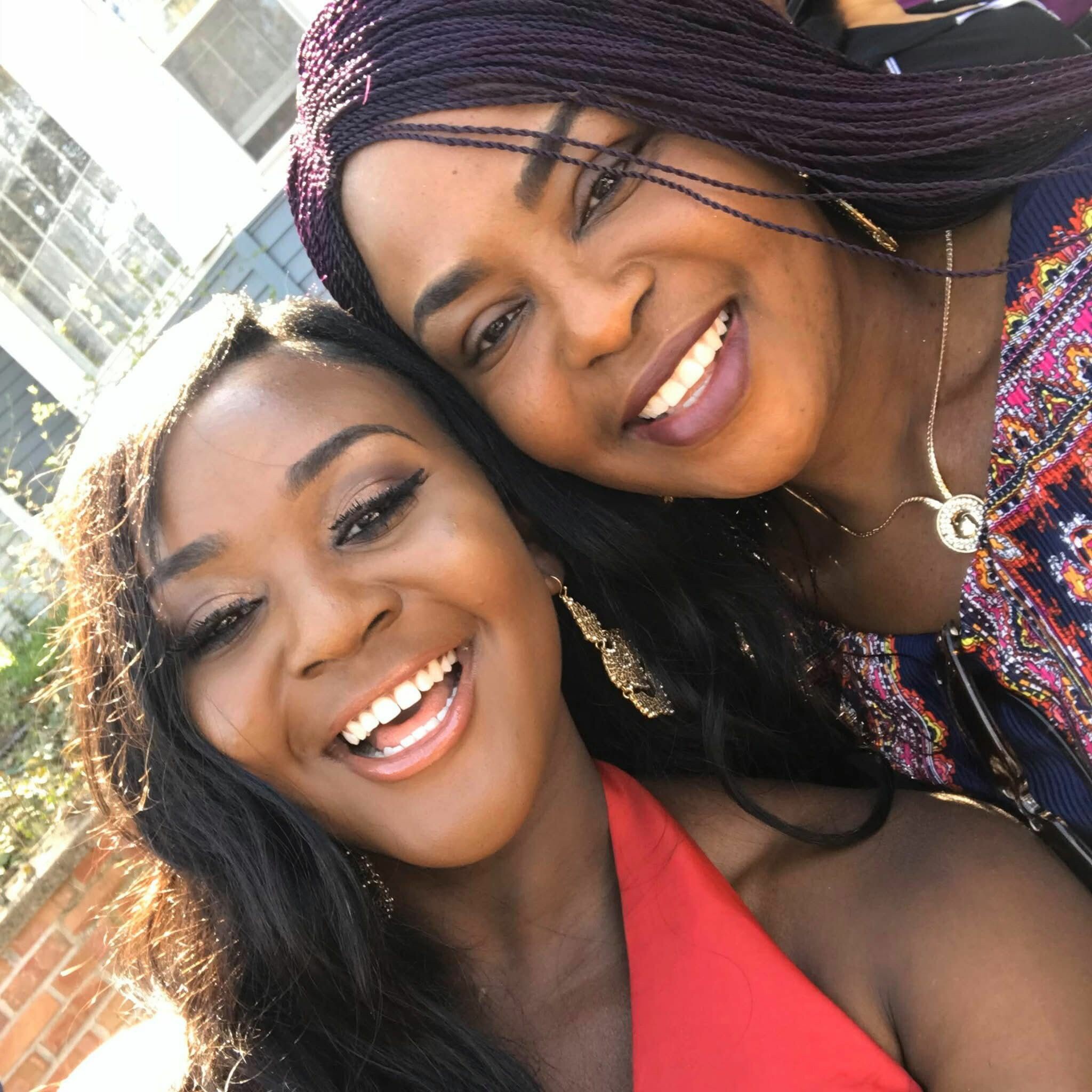 Two women smiling together for a close-up selfie outdoors, one with braids and colorful clothing adornments.