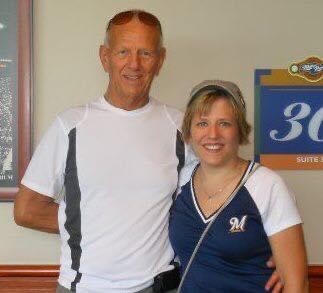 Two smiling adults posing for a photo indoors; man in a white athletic shirt with sunglasses on his head, woman in a blue varsity-style top.
