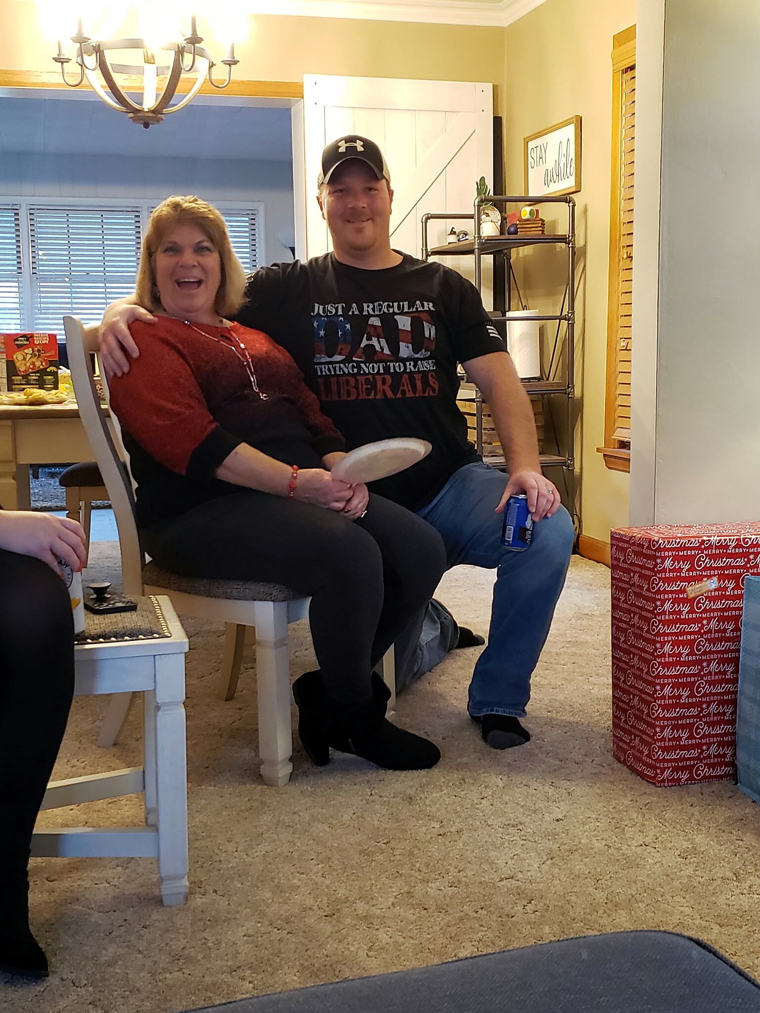 Smiling man in a black shirt and cap sits next to a smiling woman in a red sweater in a cozy living room; a large red wrapped Christmas gift sits on the floor nearby.