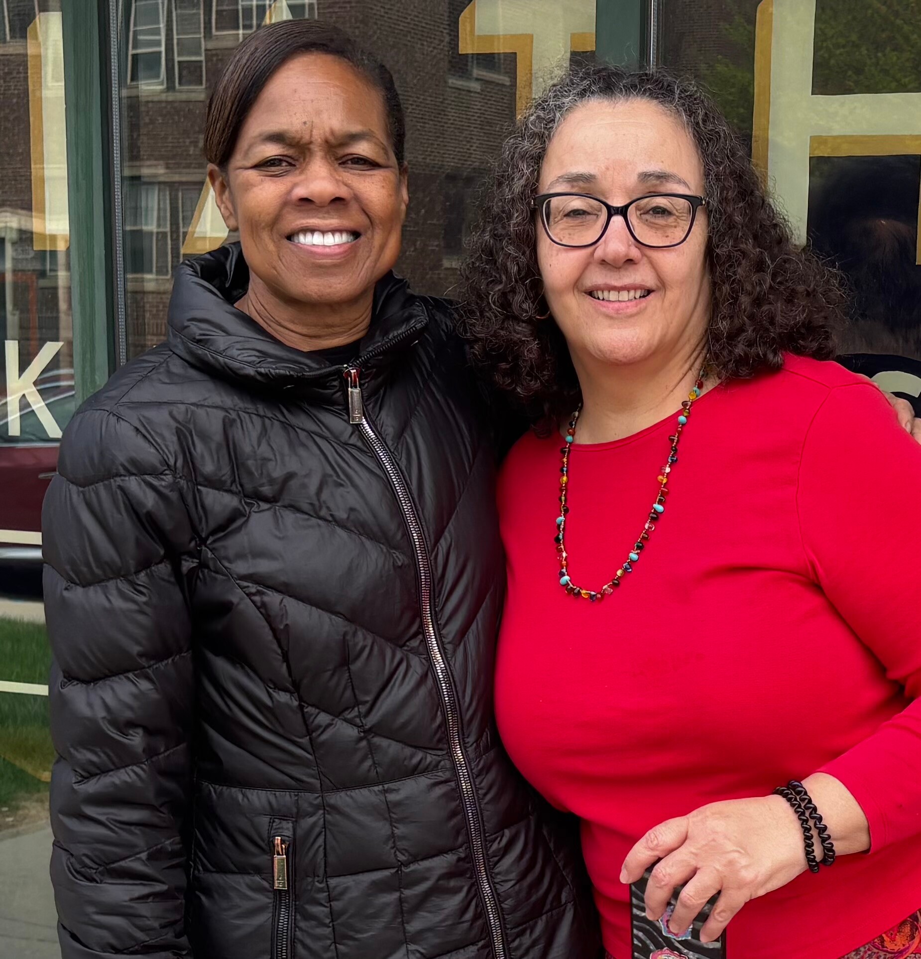 Two women stand side by side outdoors, smiling at the camera; left in a black puffer jacket, right in a red top with a colorful beaded necklace in front of a storefront window.