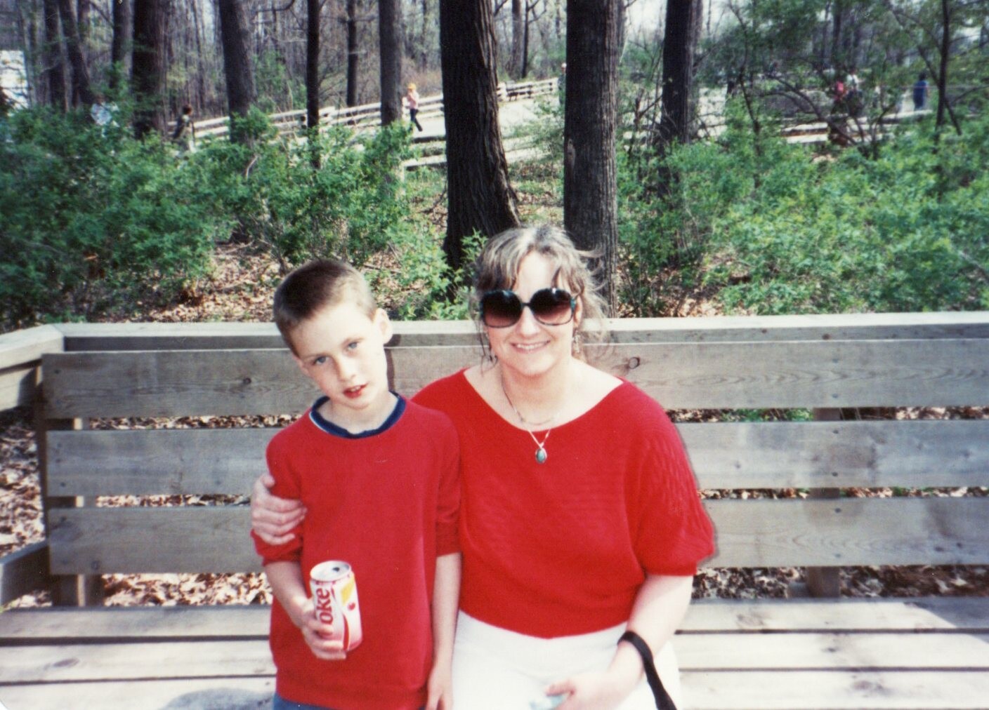Woman in red top and sunglasses sits beside a boy in a red shirt holding a Coke can on a wooden park bench, with trees in the background.