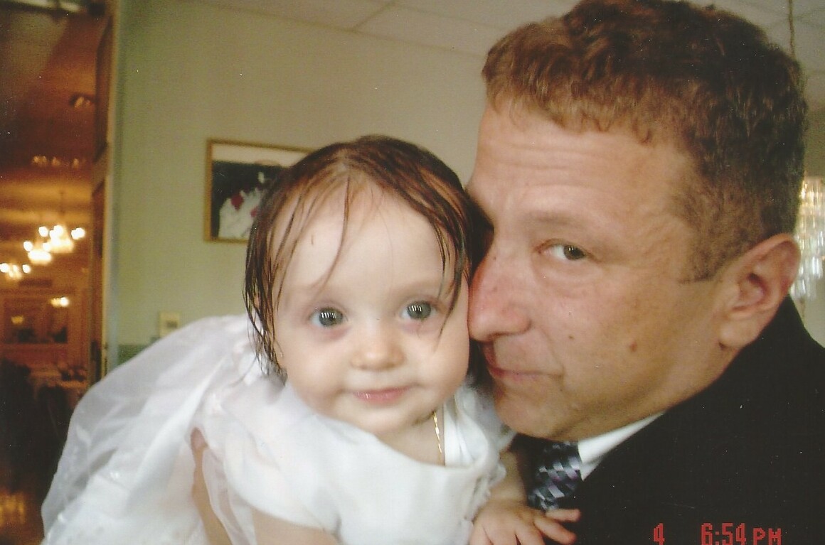 Man holding a smiling baby close to his face, the baby wearing a white dress and looking at the camera, indoors.