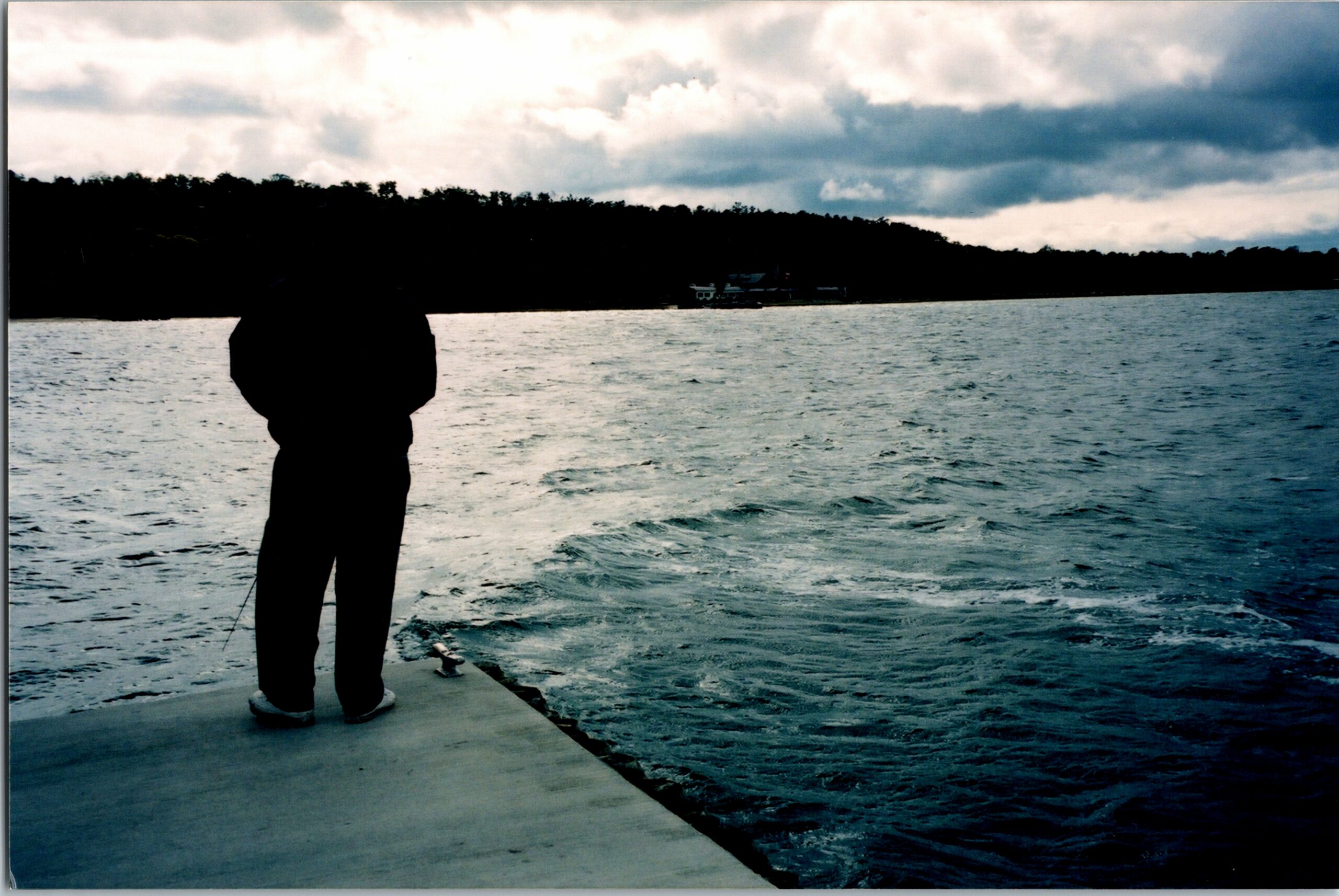 Silhouette of a person standing on a pier, looking over a choppy lake toward a tree-lined shore and cloudy sky.