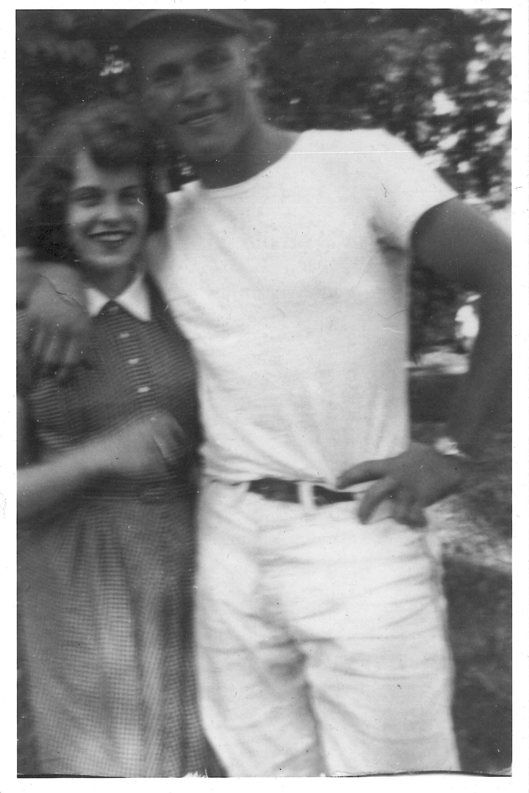 Two friends posing outdoors, smiling at the camera; left person in a checkered jacket, right person in a light shirt with trees in the background.