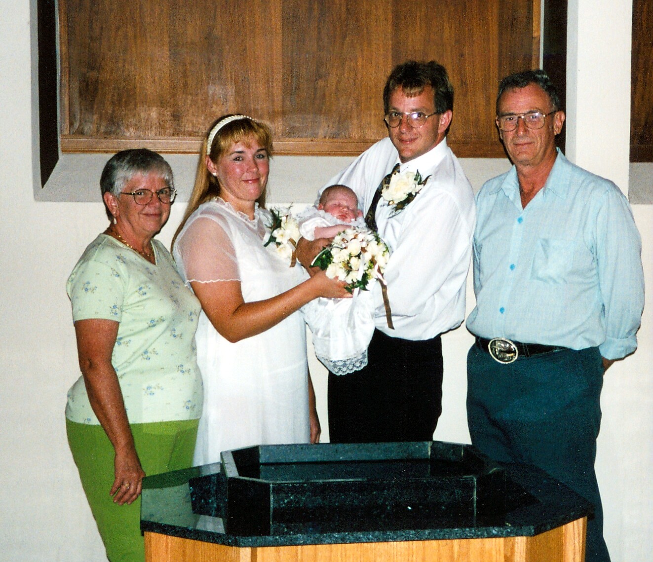 Bride and groom cradle a newborn in white, flanked by two older relatives at a baptism/wamily portrait setting.