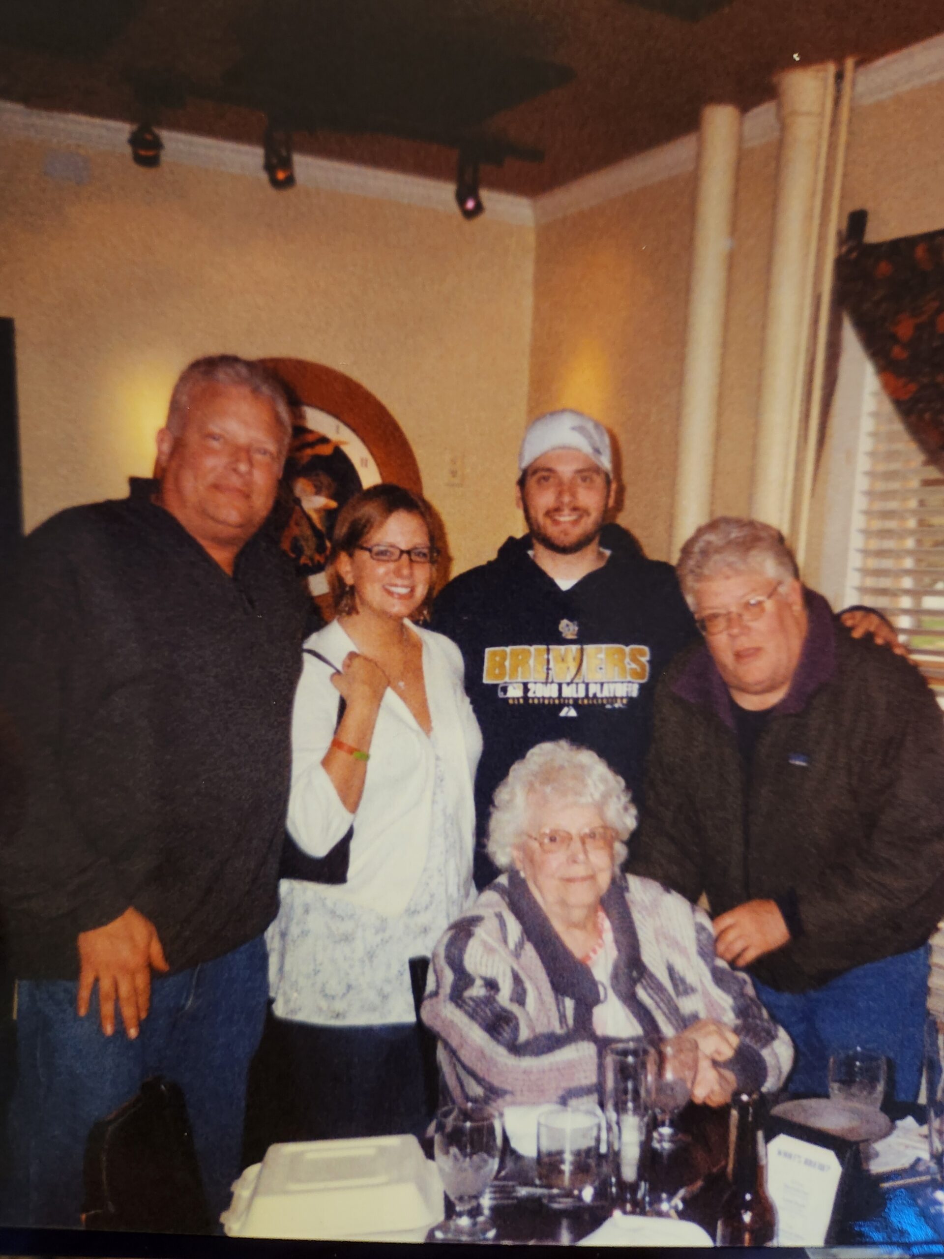 Five adults posing together at a restaurant table, smiling, with an elderly woman seated in front of them at the dinner table.