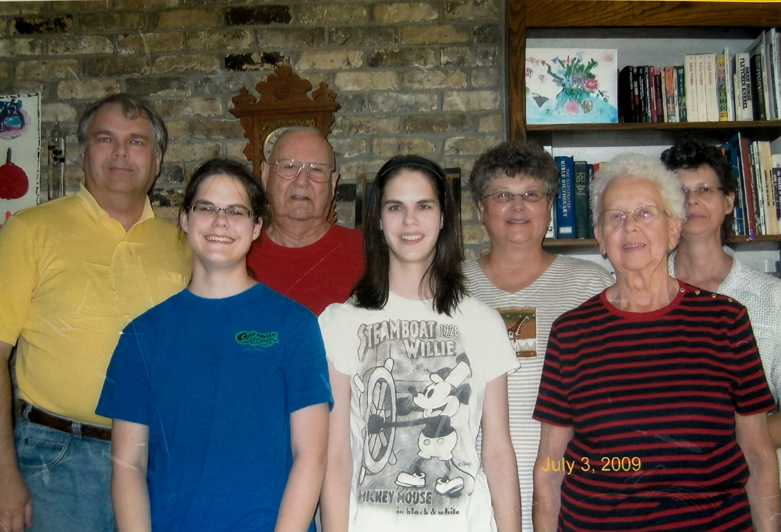 Group of seven people posing together indoors in front of a brick wall and a bookshelf, smiling for a family photo (date stamp July 3, 2009 visible).