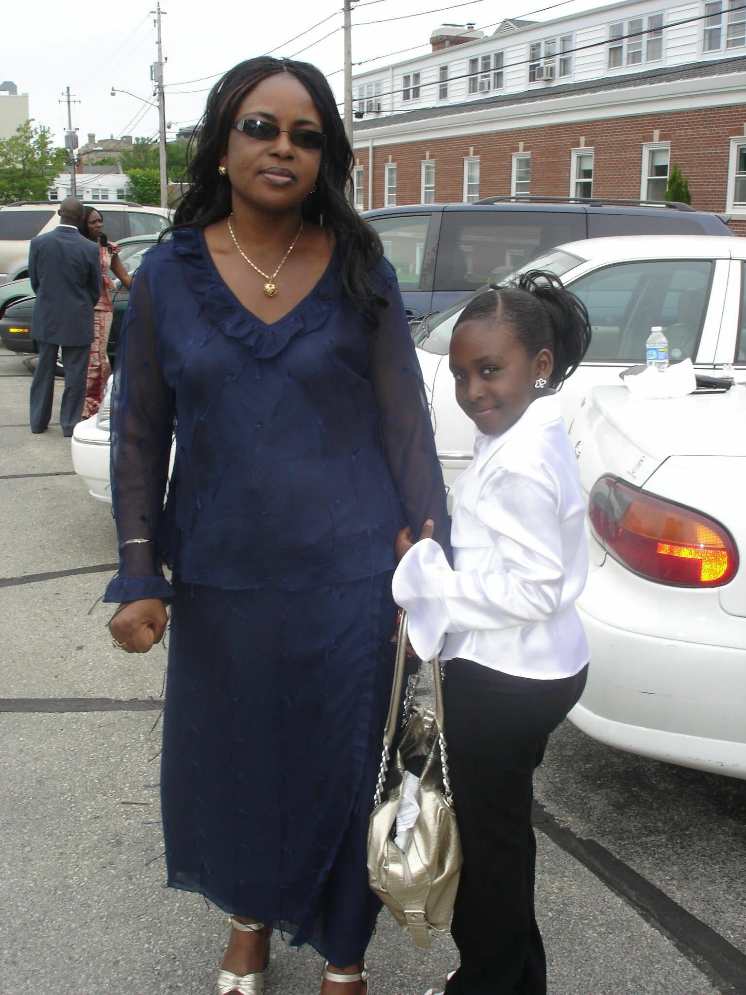 Two women posing on a city street: a tall woman in a navy blue dress with sunglasses and gold necklace, and a girl in a white blouse with a gold handbag beside her.