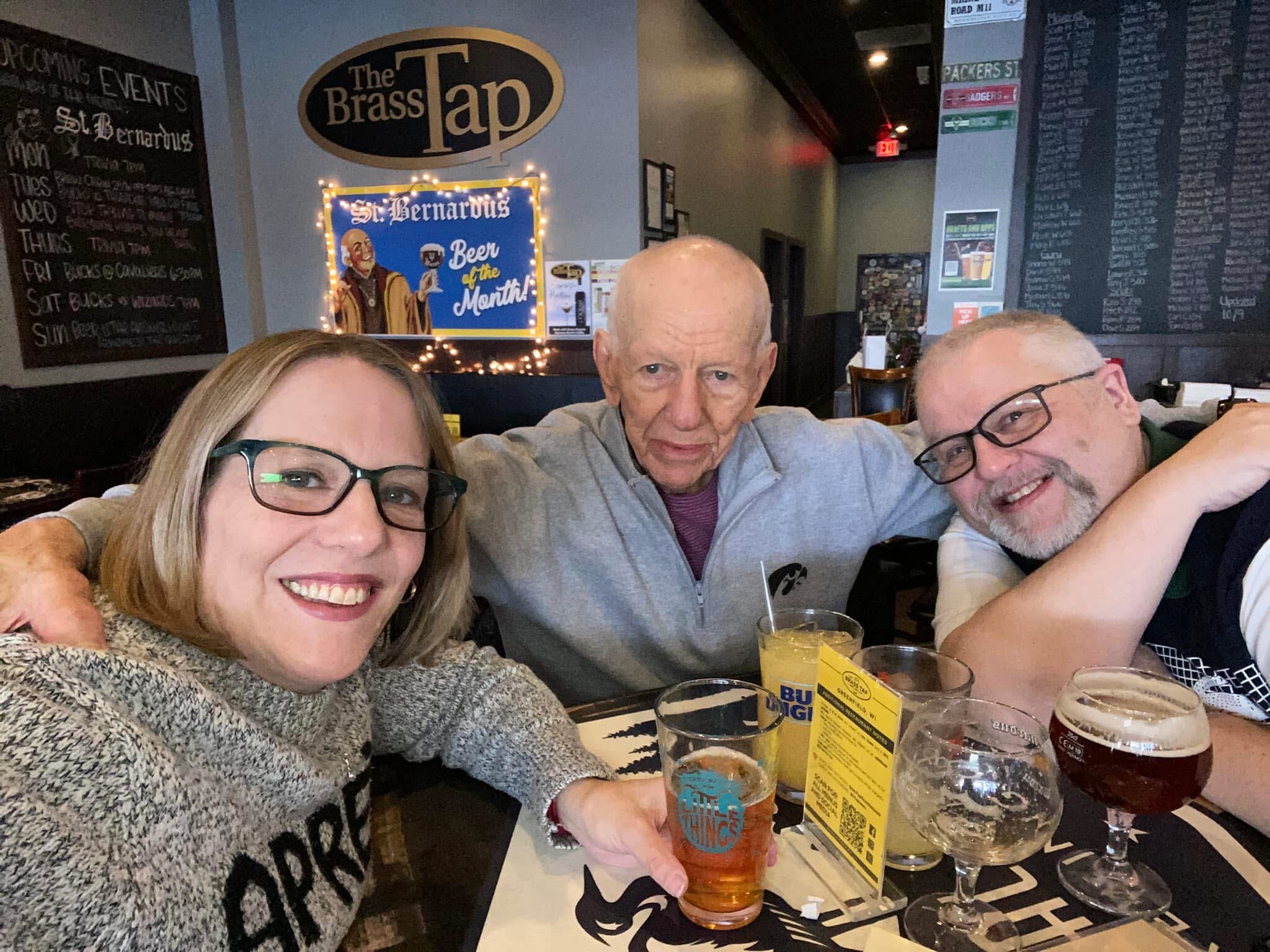Three friends take a cheerful selfie at a Brasserie-style bar; beers and glasses on the table, and a 'The Brass Tap' sign in the background.