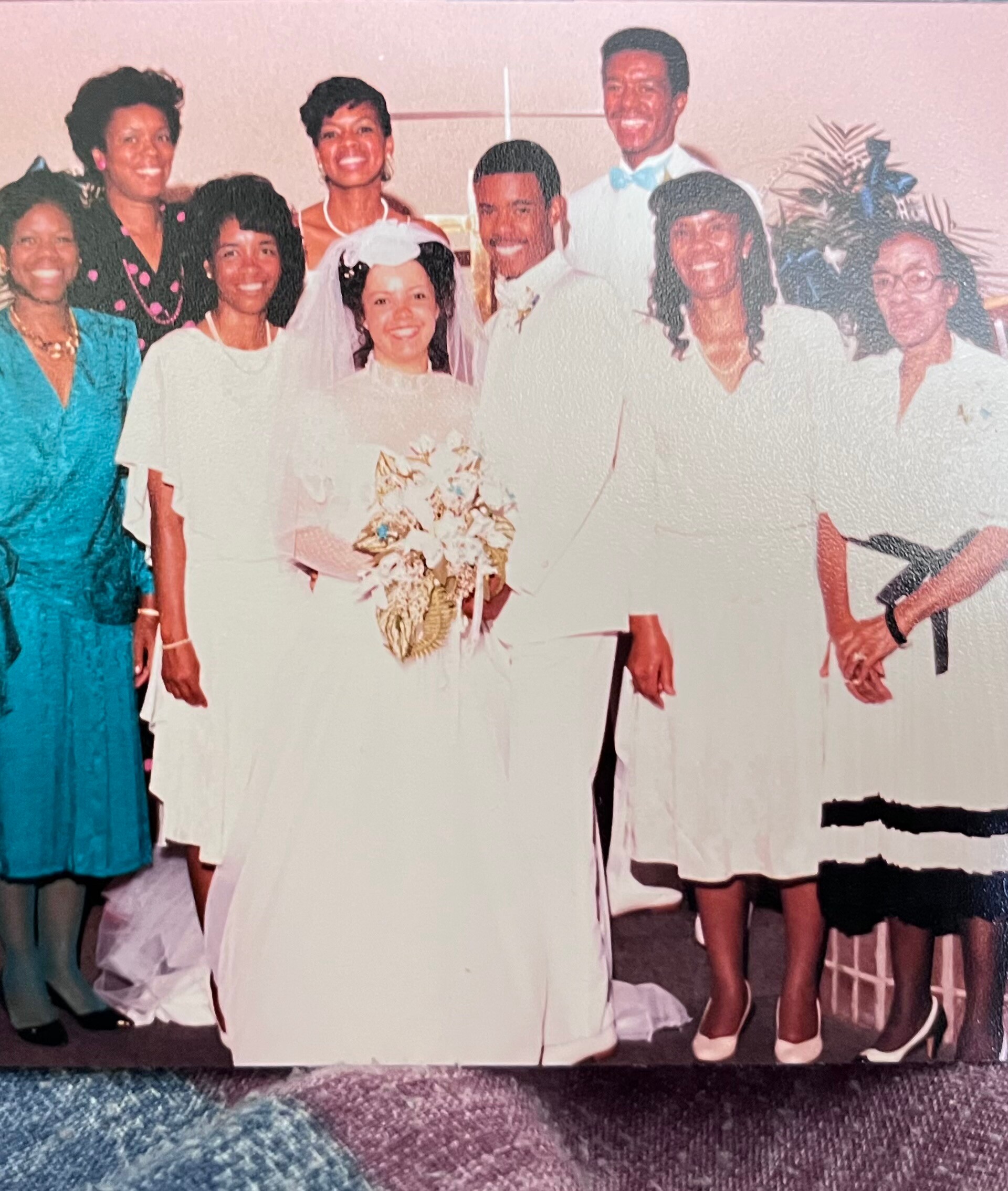 Wedding group photo: bride in white gown and veil with groom, surrounded by friends and family at a celebration.