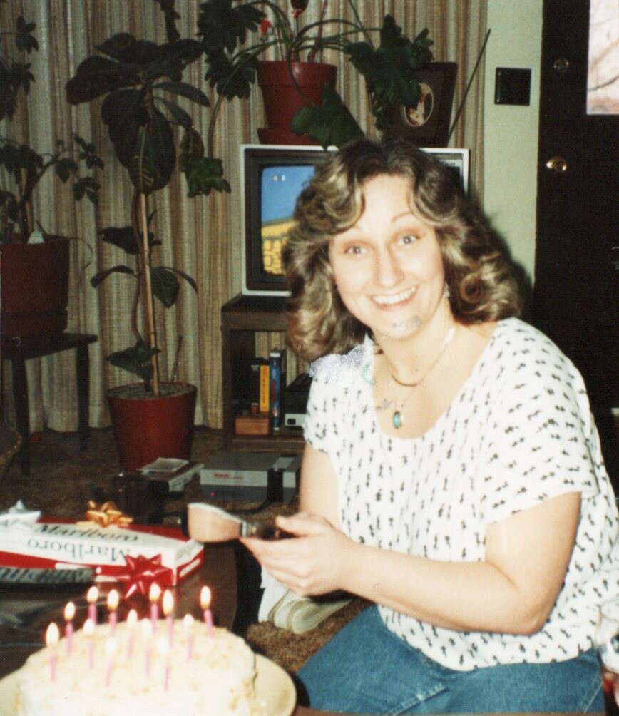 Woman smiling and holding a knife beside a birthday cake with lit candles in a living room.