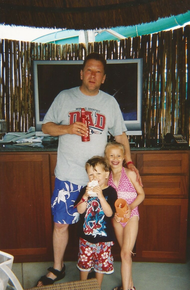 Man in a gray t-shirt and blue floral shorts stands behind two kids, holding a red bottle, in a sunny backyard.