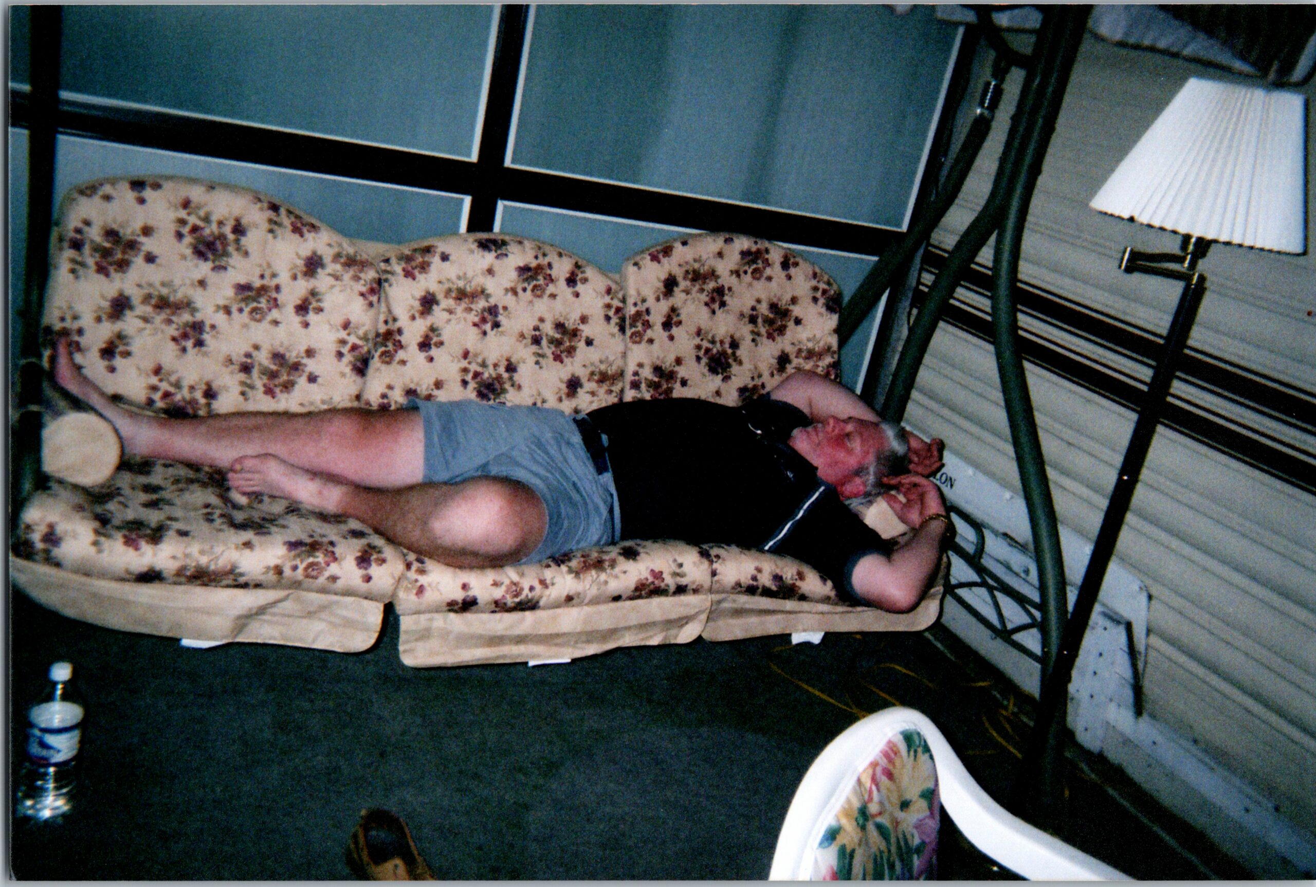 Man lying on a beige floral couch with his head in his hands in a cluttered room, a lamp and cords nearby.