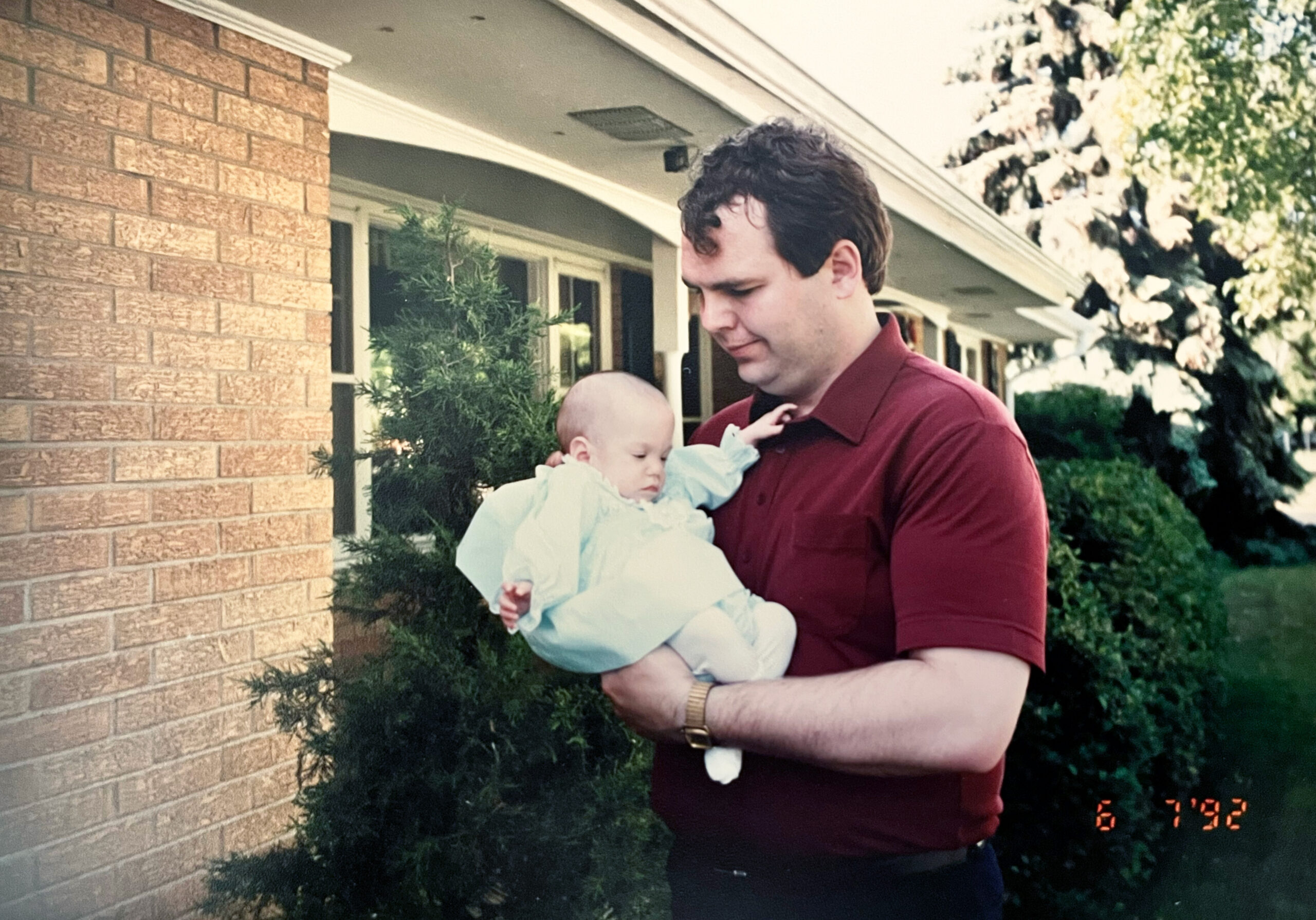 Man in a maroon shirt holding a baby in a light blue outfit outside a brick house with bushes nearby.
