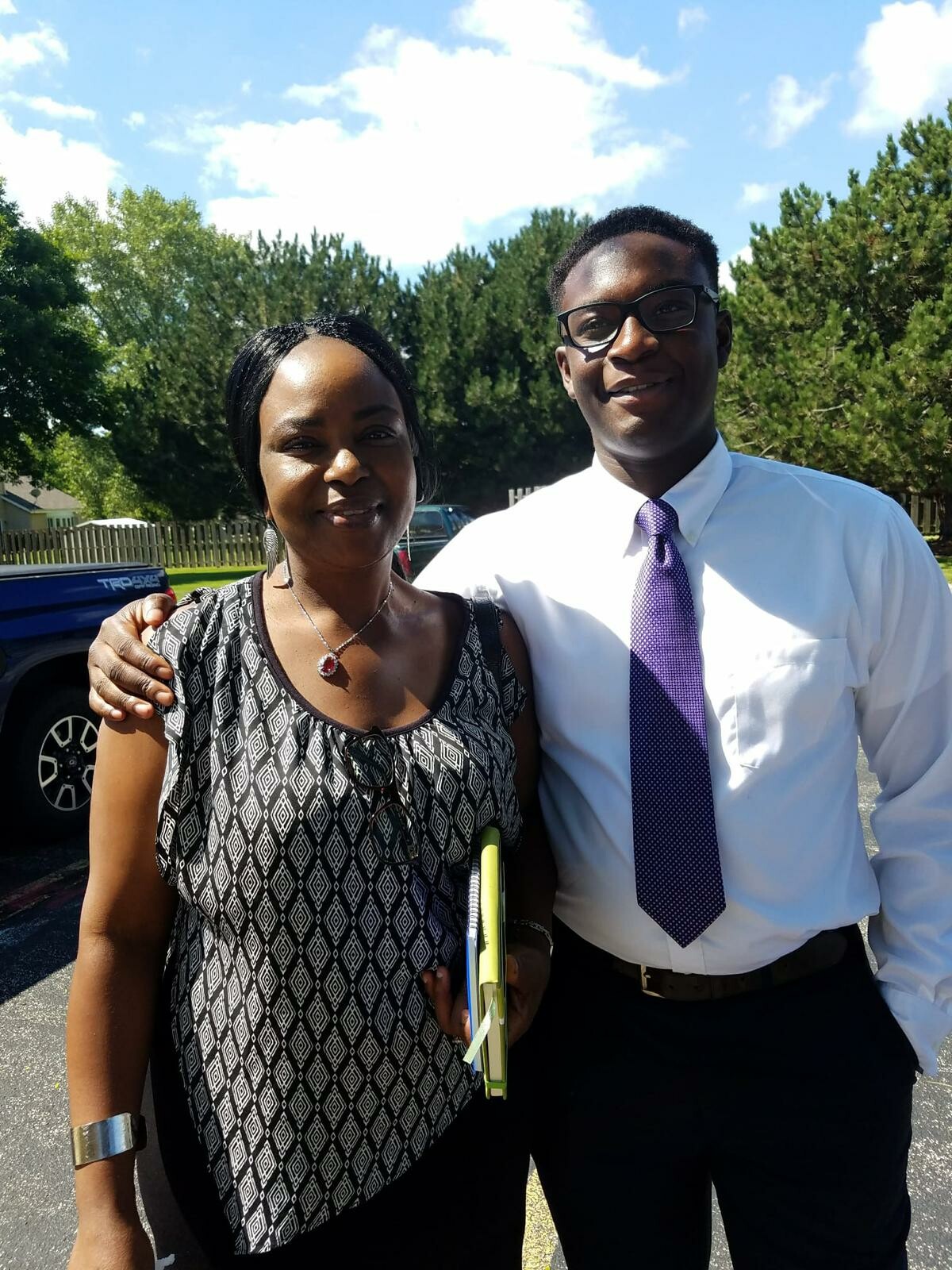 Two smiling adults posing together outdoors in a parking lot, with trees and blue sky in the background; the woman wears a patterned black-and-white top and necklace, holding a folder, and the man wears a white shirt with a purple tie and glasses, arm around her shoulder.