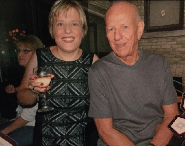 Smiling woman in a geometric black-and-white dress holds a cocktail beside an older man in a gray shirt at a party stack of friends in the background
