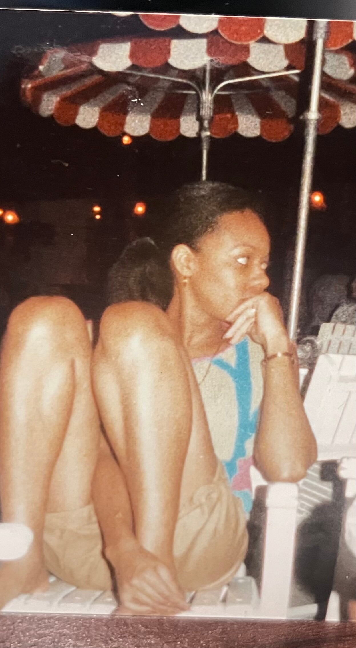 Woman sitting cross-legged on a bench at night, under a red-and-white balloon canopy, resting her chin on her hand.