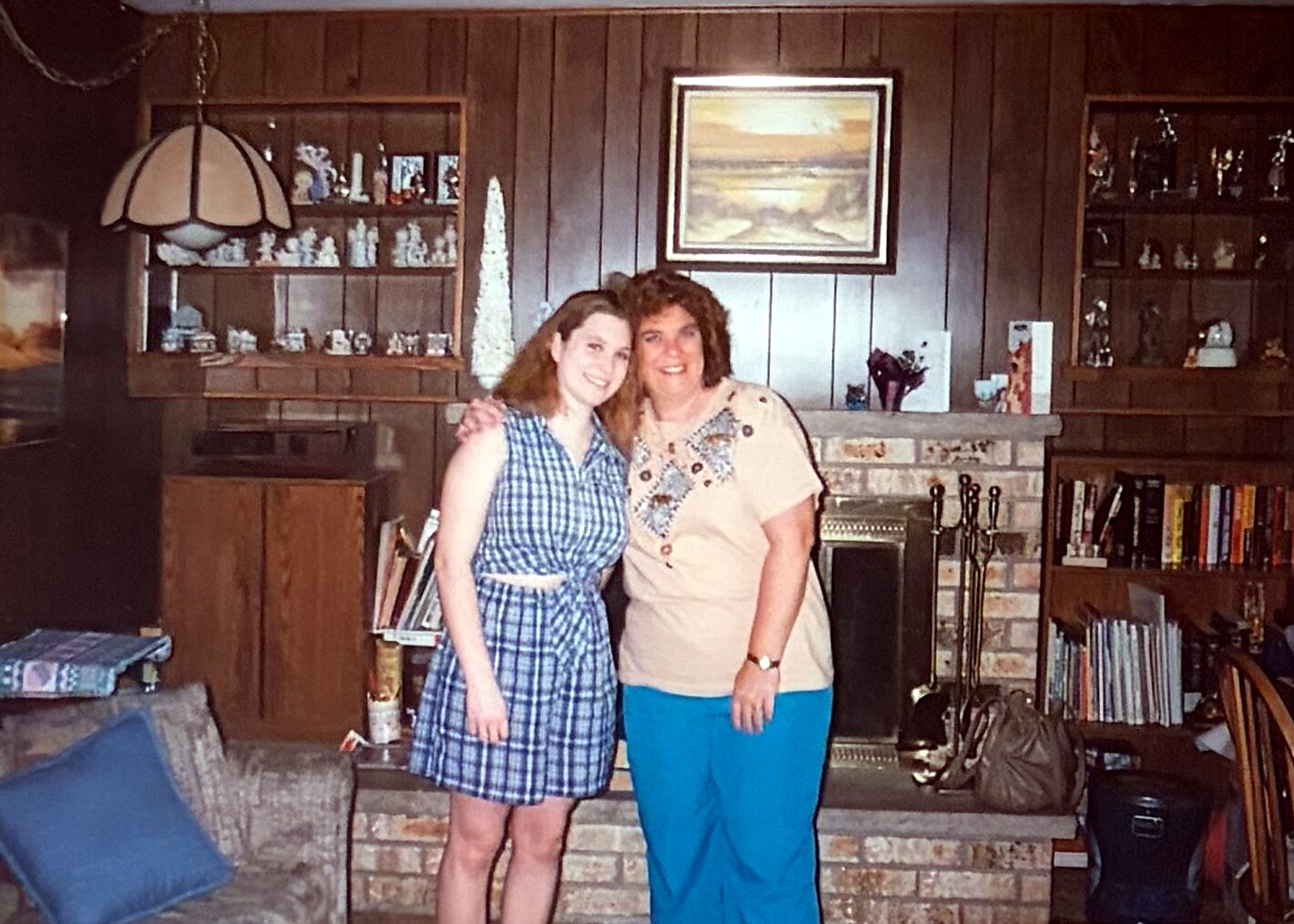 Two women stand arm-in-arm and smile at the camera in a wood-paneled living room with a brick fireplace and decorative shelves behind them.