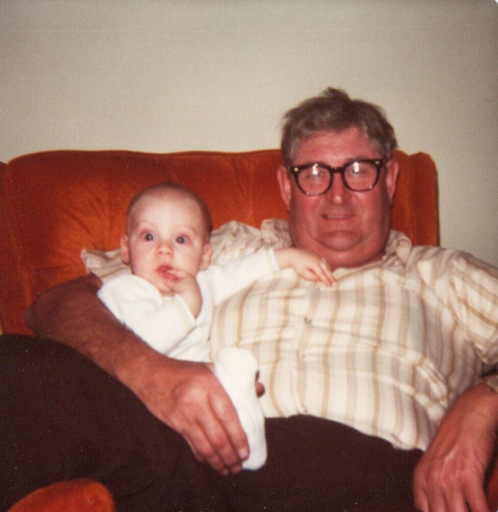 Older man with glasses sitting on an orange couch, holding a baby on his lap.