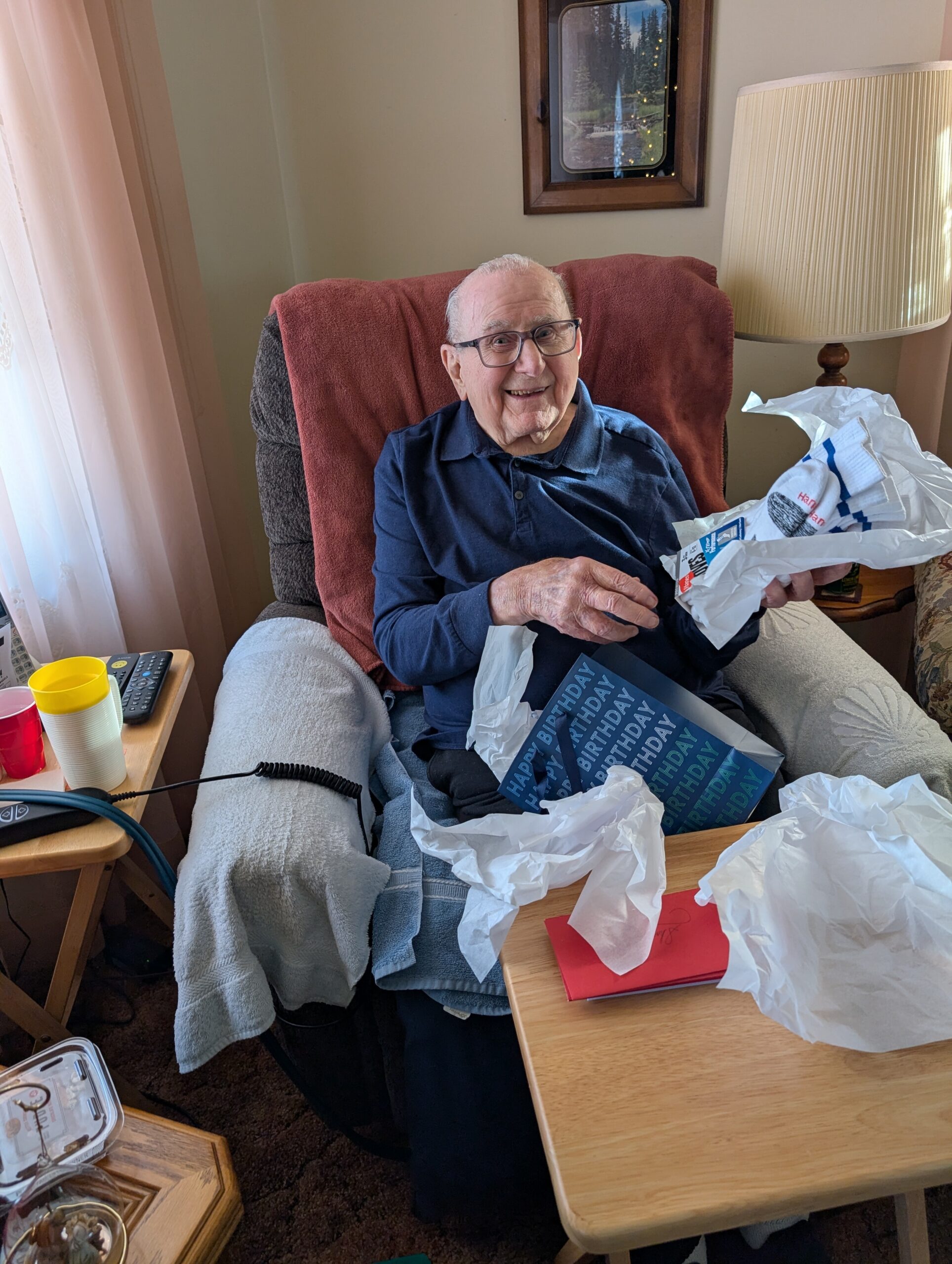 Smiling elderly man in glasses sits in a recliner with a red blanket, holding birthday cards and tissue paper.