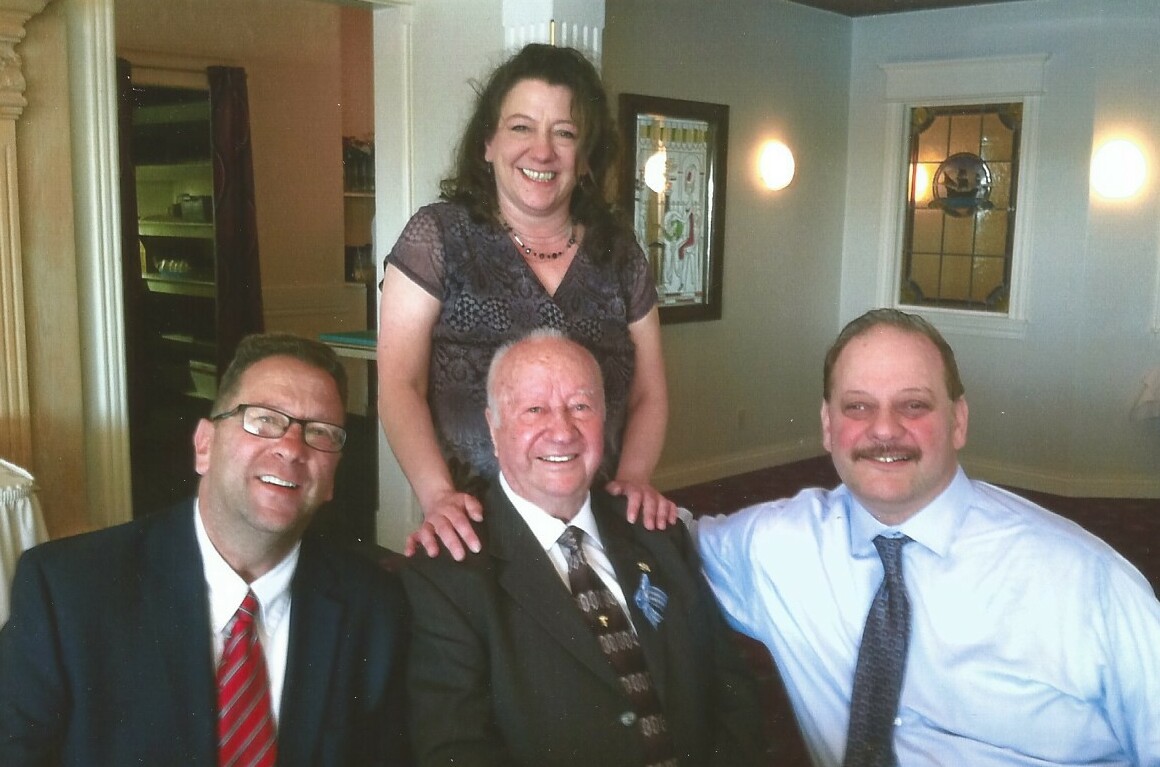 Four adults posing for a group photo indoors, all smiling, with a woman standing behind the two seated men in a formal setting.