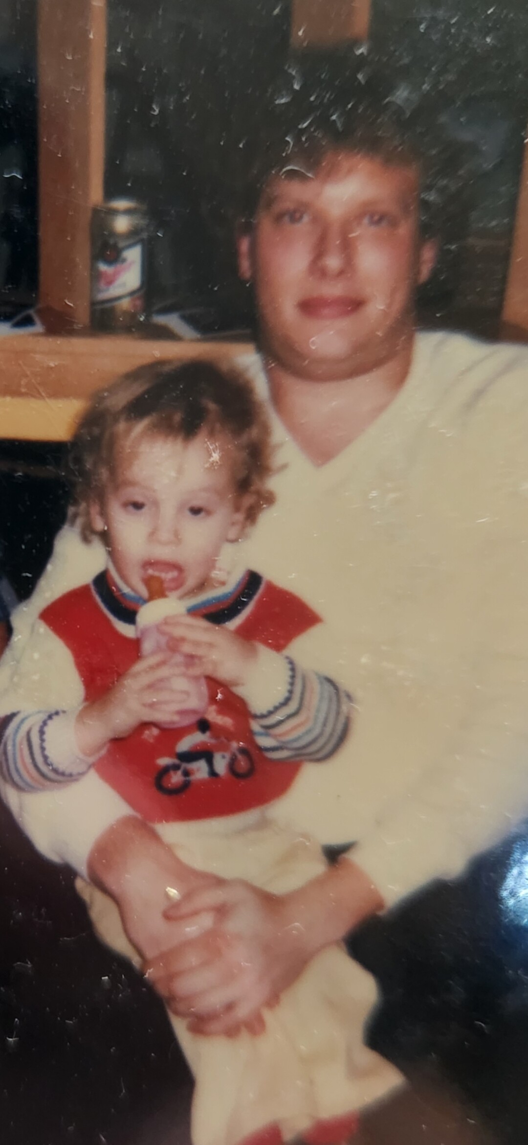 Man in a light yellow sweater holds a toddler who is drinking from a bottle.