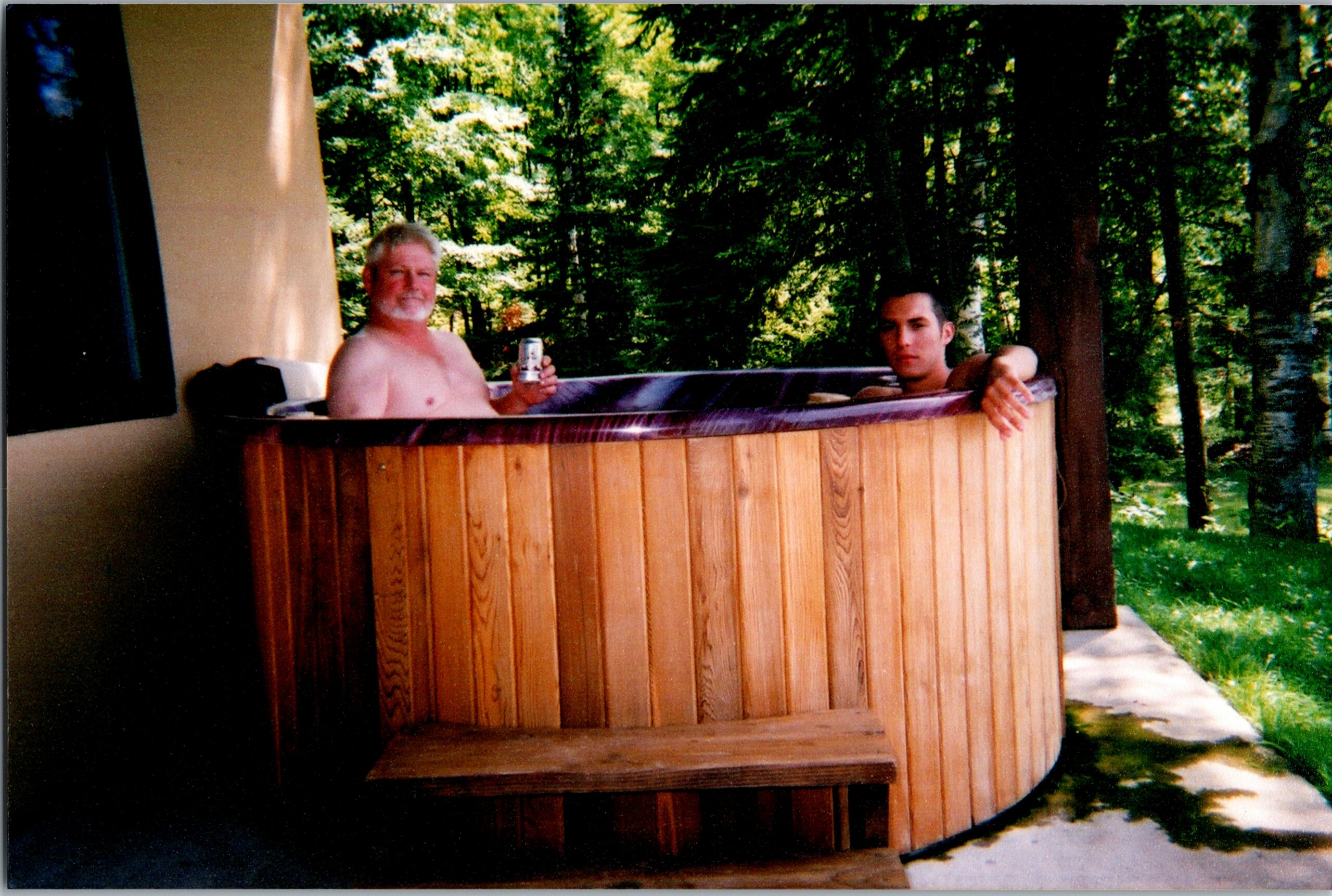 Two shirtless men soak in a wooden outdoor hot tub on a covered porch, one holding a can while forest surrounds them.