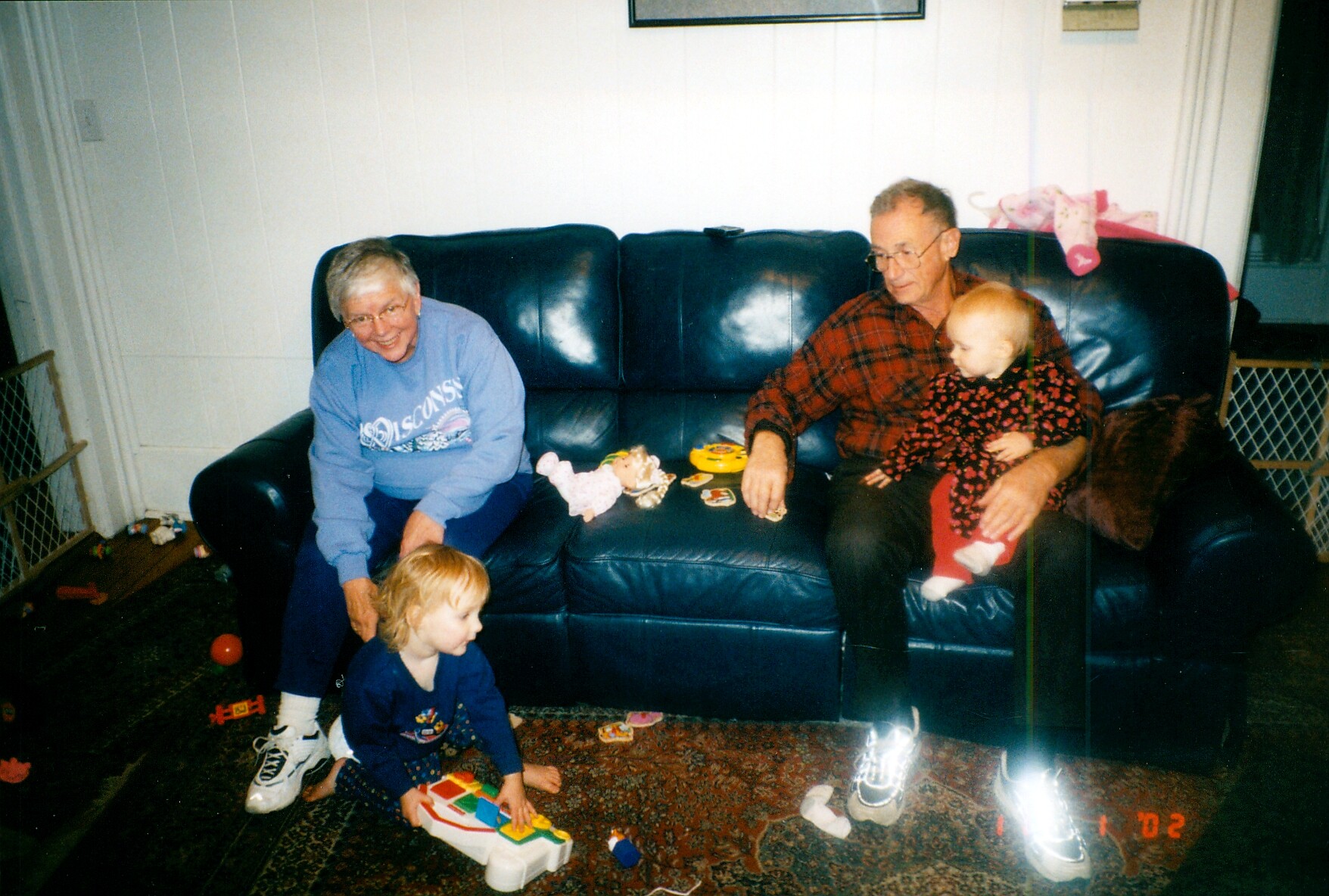 Two grandparents sit on a blue leather couch as a toddler and baby play with toys on the rug in the living room.