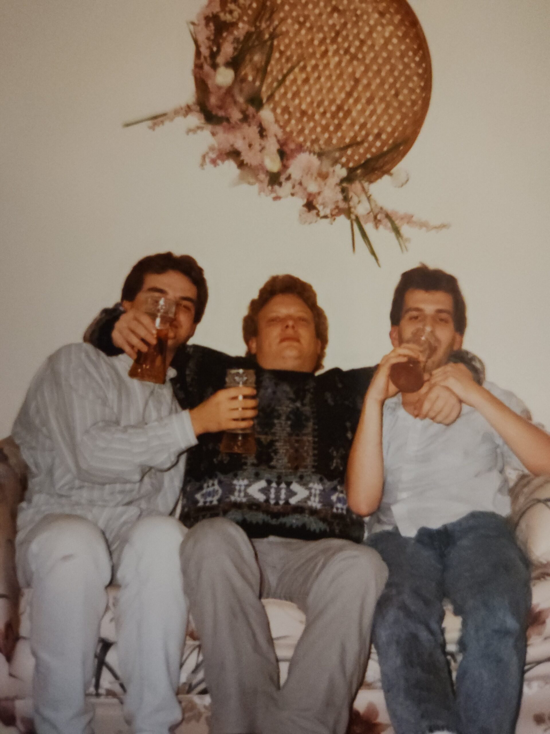 Three men sit on a couch, toasting with beer glasses, smiling, with a large floral arrangement hanging above them.