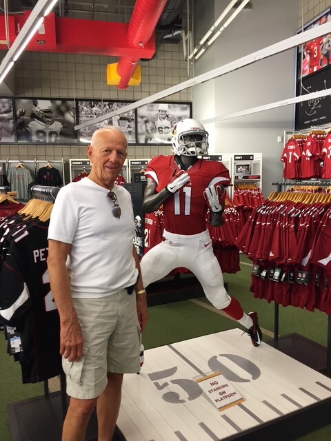Older man in white shirt and shorts posing beside a large football mannequin in a sports store display area.