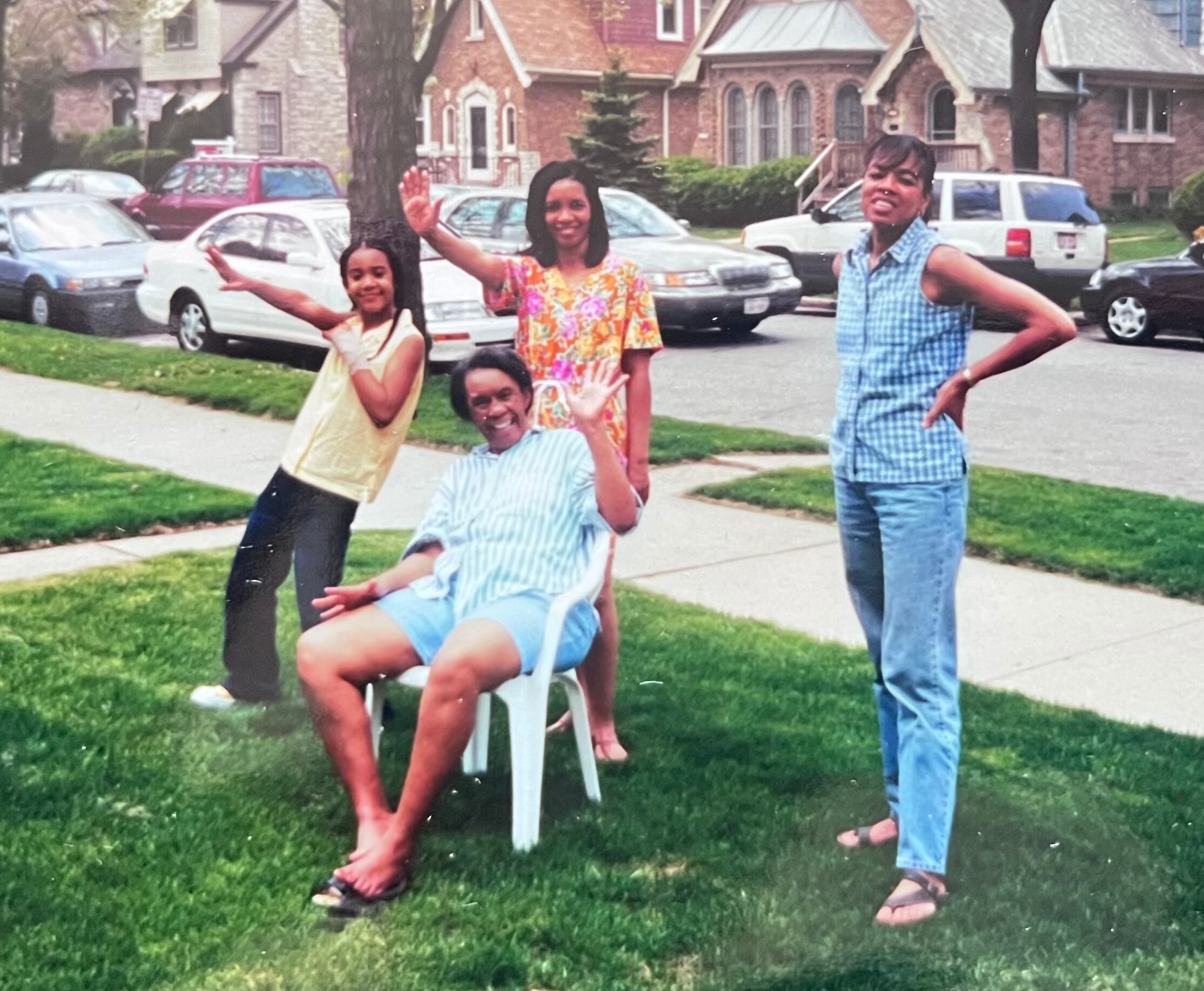 Group of friends/family posing in a yard on a sunny day, waving at the camera on a suburban street.