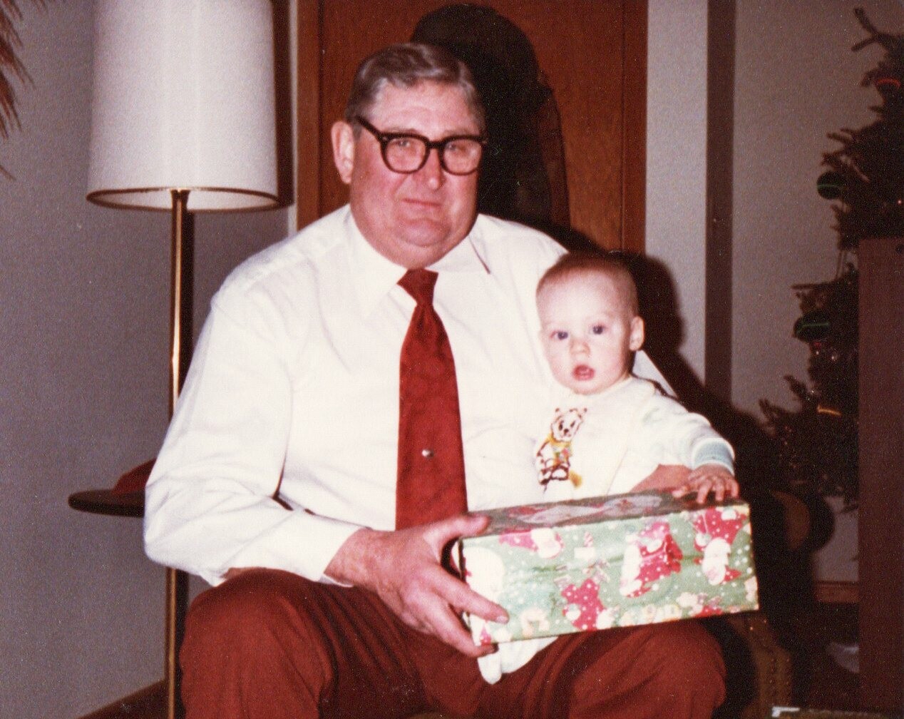 Older man with glasses sits with a baby on his lap, holding a wrapped Christmas gift box in a holiday scene.