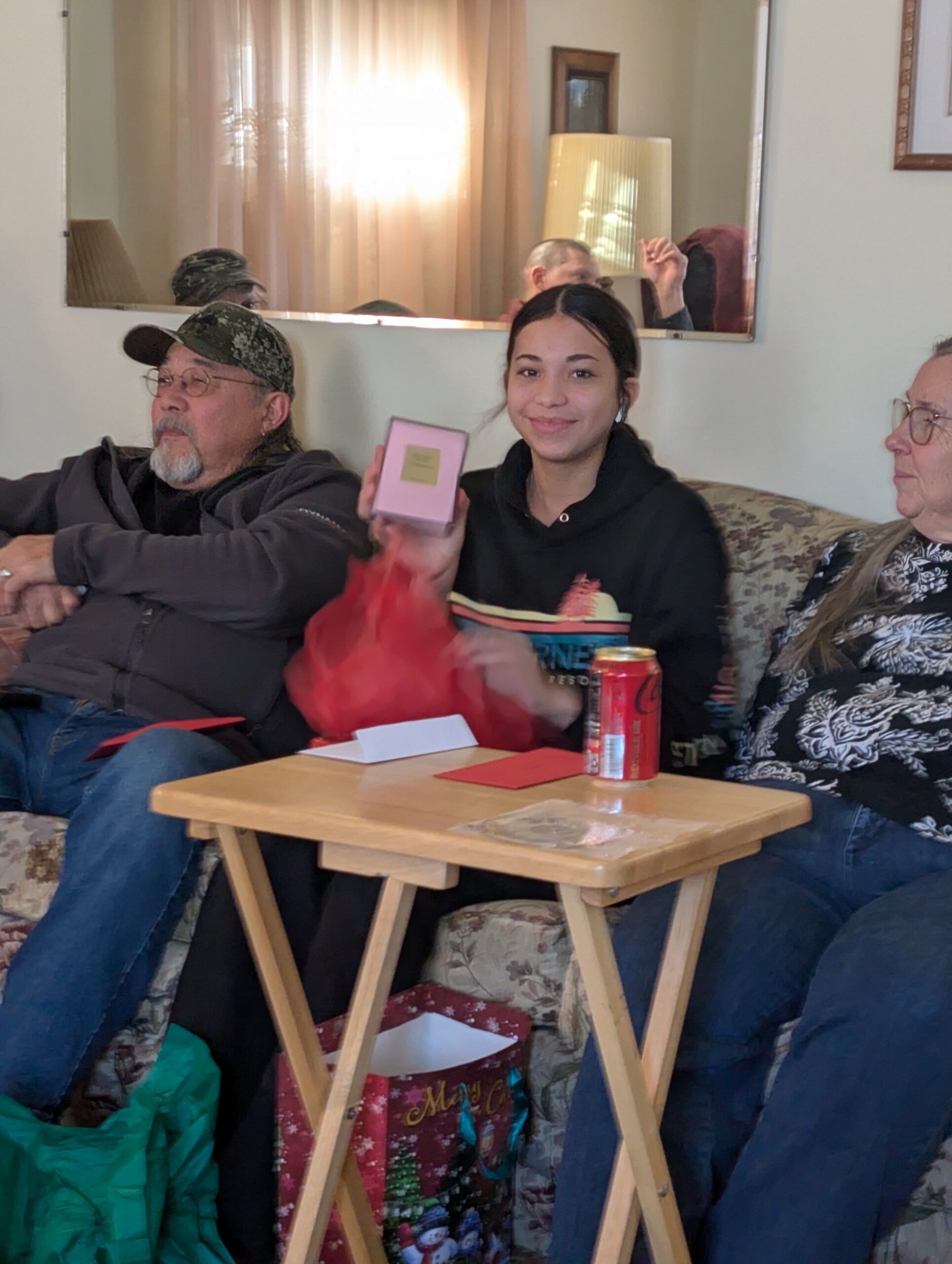 Group of three people sitting on a floral couch; a young woman in the middle holds a small pink gift box and smiles, with a red can on the table nearby.