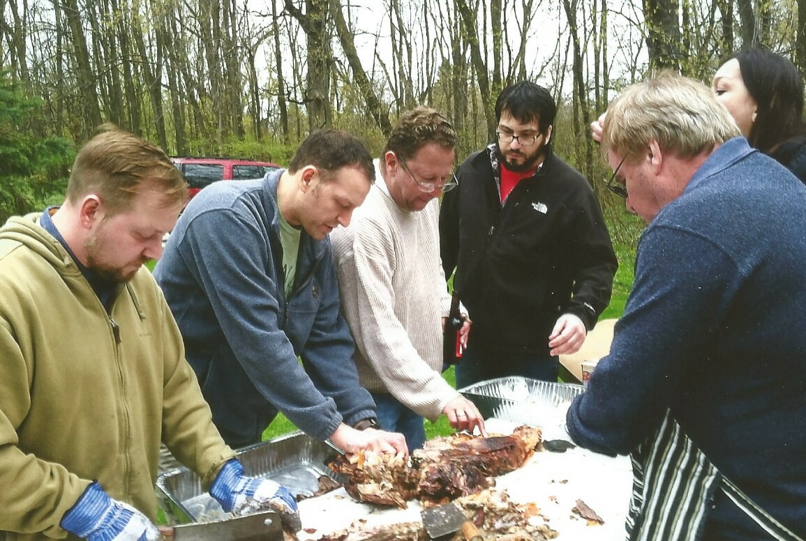 Group of men outdoors carving a large roast meat on a foil tray. They lean in, using forks and hands to separate the meat.